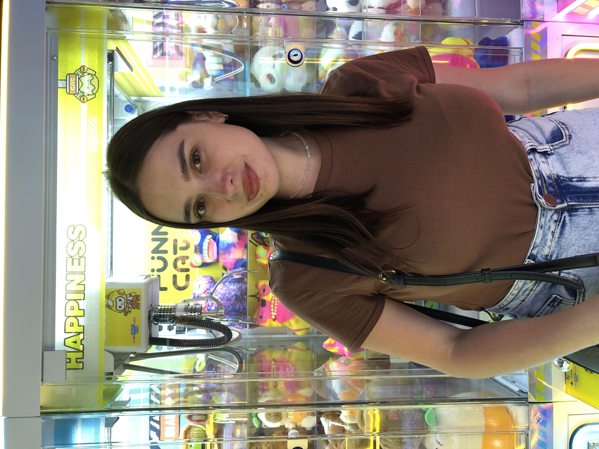 The image shows a young woman posing for a selfie in front of a claw machine. She is wearing a brown t-shirt and blue denim jeans. The claw machine is filled with various stuffed animals and toys, with bright lights illuminating the interior. The text "HAPPINESS" is prominently displayed in yellow lettering on the side of the machine. There is also a smaller graphic of a cartoon character with the text "CHEN TIAN" below it. The woman has long dark hair and is looking directly at the camera with a slight smile. The scene appears to be indoors, possibly at an arcade or entertainment center. The lighting is bright and colorful, typical of such establishments. The woman's posture and expression suggest a lighthearted and casual mood. There are no clear indicators of the time of day or weather conditions. The location is identified as Chaikovsky, Russia.