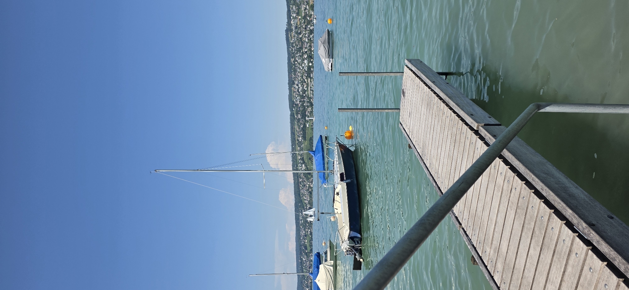 The image captures a serene scene on the lake of Zurich in Oberrieden, Switzerland, on a clear, sunny day. In the foreground, a wooden pier with metal railings extends into the water, suggesting a place for docking or recreation. The water is a tranquil turquoise, reflecting the bright blue sky dotted with a few wispy clouds.
Several sailboats are moored in the lake. One prominent sailboat in the mid-ground has a blue cover over part of its deck and a tall mast. To its left, another sailboat is partially visible with a beige cover. Further in the distance, more sailboats can be seen, some appearing smaller and further away. Another motorboat, covered with a gray tarp, is also anchored in the water. Two orange buoys are visible, marking areas in the lake.
The background reveals a picturesque landscape of Oberrieden. A densely populated hillside covered with houses and lush green vegetation rises from the lakeshore. The town appears to be nestled harmoniously within the natural environment. The overall atmosphere of the image is one of peaceful summer leisure. There are no people visible in the scene.