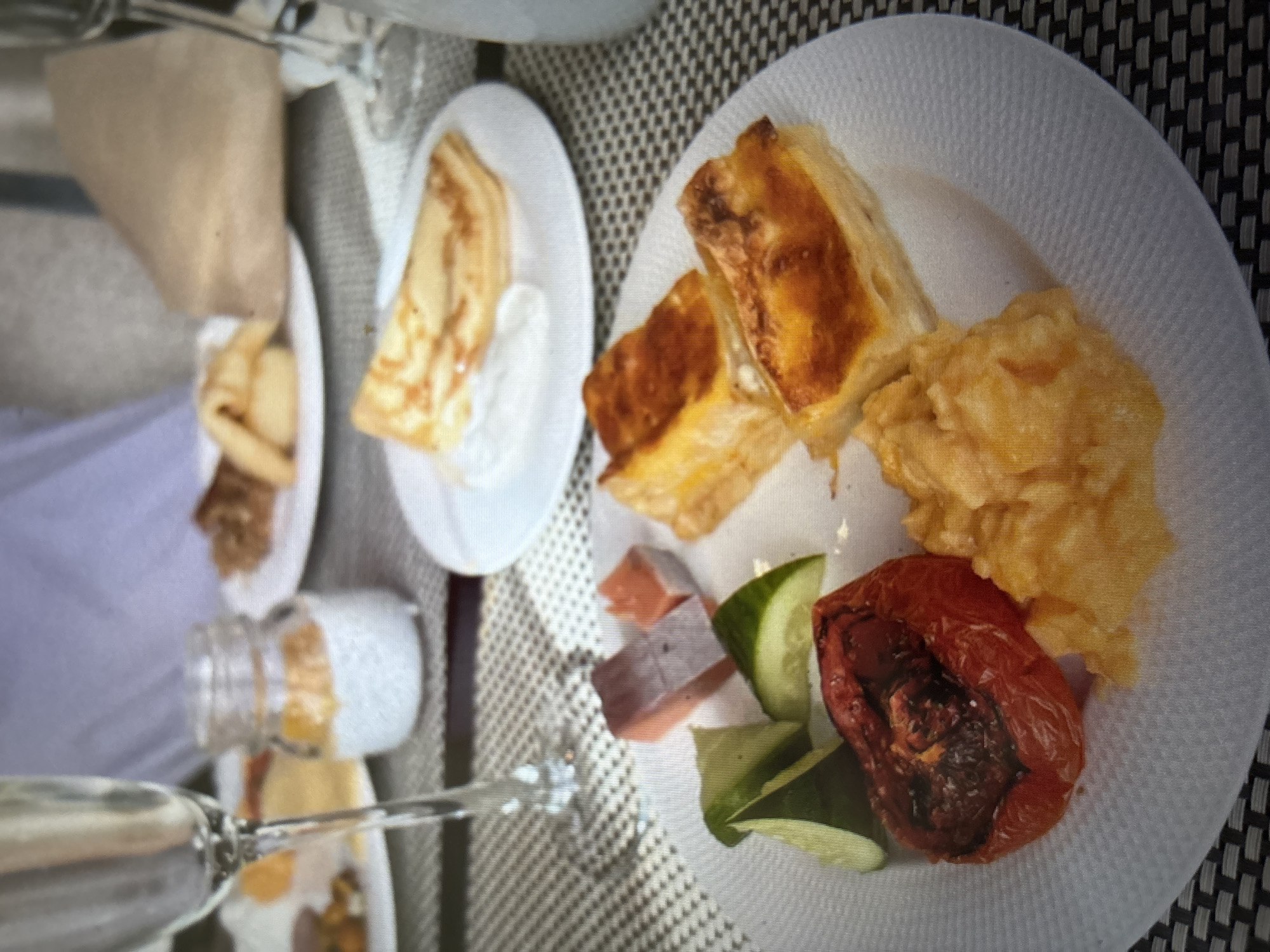 A close-up, slightly blurry overhead shot shows a table with various breakfast items. In the foreground, a white plate holds a roasted tomato, scrambled eggs, and slices of cucumber. To the right of that plate are two pieces of a pastry or quiche. Further back, a white plate contains a folded crepe with sauce. Behind that, a small jar with a white substance and orange topping sits on the table. Another white plate with pastries is visible in the background. A champagne flute is on the left side of the frame, and another glass is partially visible on the right. The table is covered with a woven placemat. The scene appears to be indoors, possibly at a restaurant. The specific location in Novosibirsk, Russia, cannot be determined from the image.