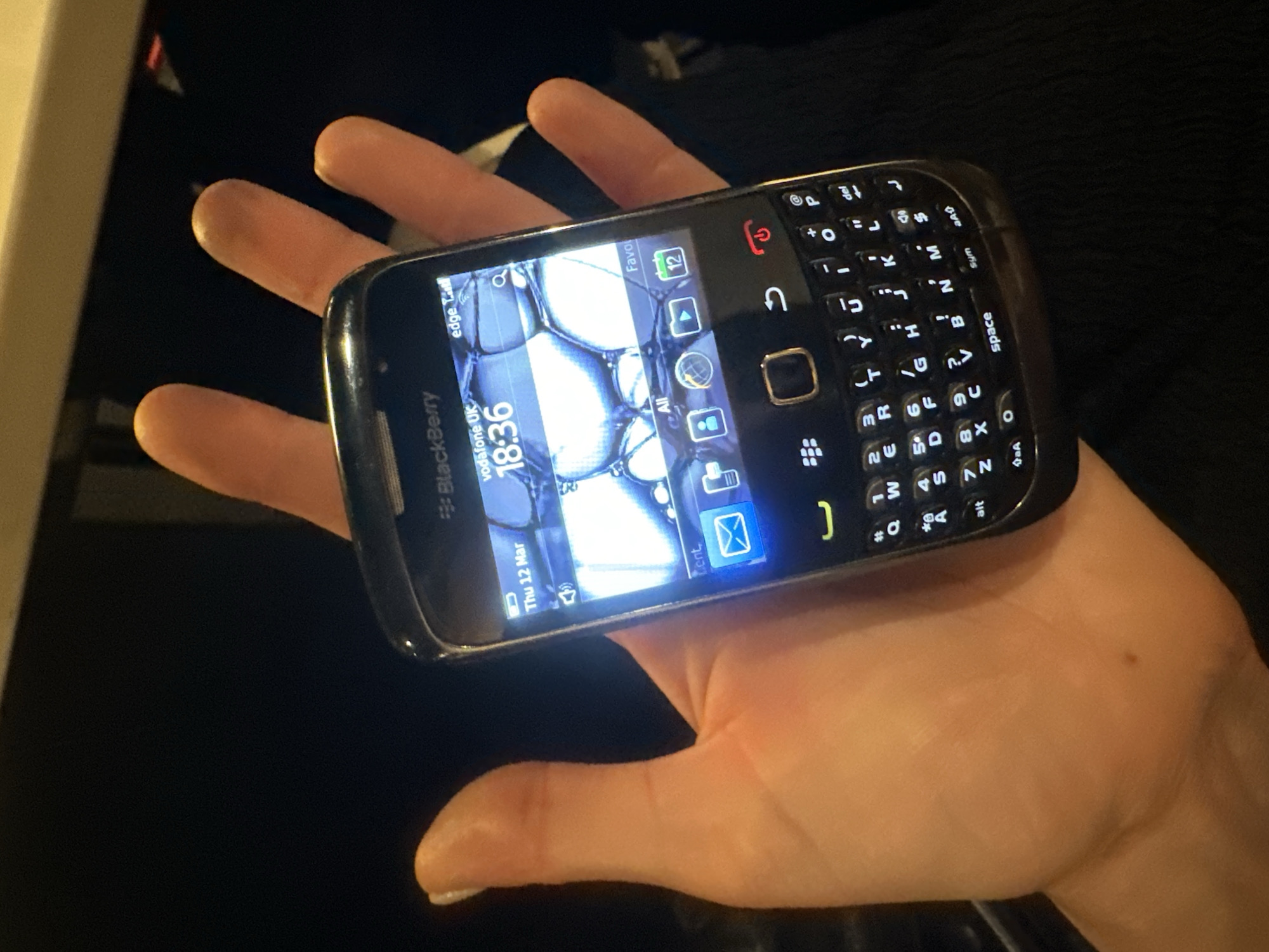 A human left hand holds a black BlackBerry smartphone in a vertical orientation within a dimly lit indoor environment. The phone's illuminated screen displays "Thu 12 Mar" and "18:36." The network carrier is identified as "vodafone UK," operating on "edge" with full signal strength. The screen wallpaper features a blue-toned abstract pattern of bright, interconnected circular forms. An application dock is visible below the time, with an envelope icon highlighted in blue. The smartphone features a full QWERTY keyboard with white backlighting, an optical trackpad, and a red illuminated end-call button. The hand exhibits fair skin with a small, dark mole near the base of the thumb and a white-manicured thumbnail on the left thumb. The background consists of dark, out-of-focus elements, with vague lighter shapes in the upper frame and dark clothing visible in the lower right. The phone's network connection indicates operation within the United Kingdom, consistent with the stated location of London, United Kingdom.