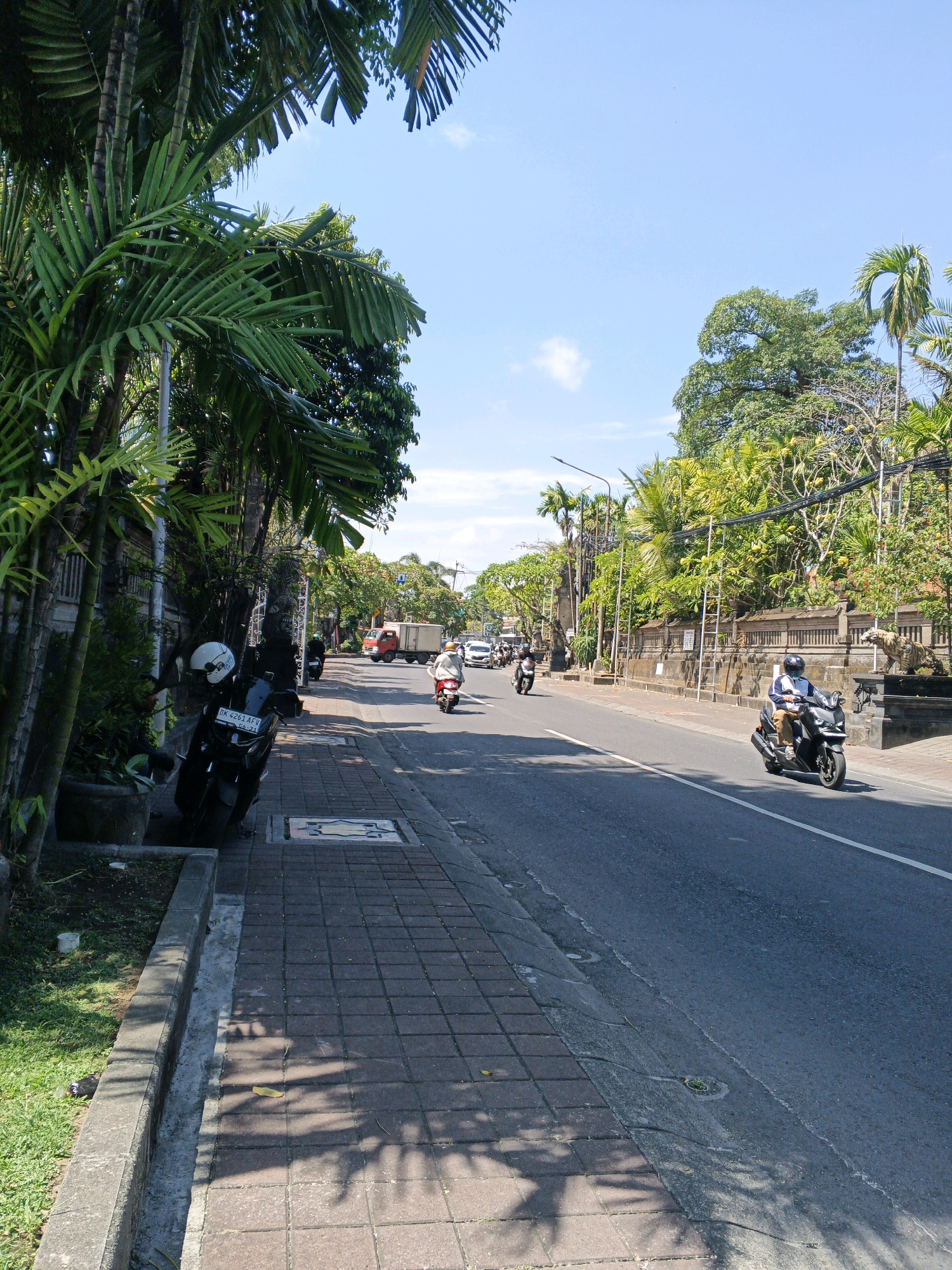 The image depicts a sunlit street scene in North Kuta, Indonesia. A paved sidewalk runs along the left side of the road, bordered by a low concrete curb and a line of lush green vegetation, including palm trees and potted plants. Several parked motorcycles are visible on the sidewalk, one of which has a white helmet on its seat and a license plate reading "DK 4261 AFV." The street itself is asphalt and appears to be a multi-lane road with moderate traffic. In the distance, a red truck is visible, along with several other vehicles and motorcycles. Pedestrians and motorcyclists are present on the street and sidewalk. The buildings lining the street are partially obscured by dense foliage and trees, with a stone wall and decorative elements, including what appears to be a tiger statue, visible on the right side of the road. The sky is clear and blue, indicating a bright day.