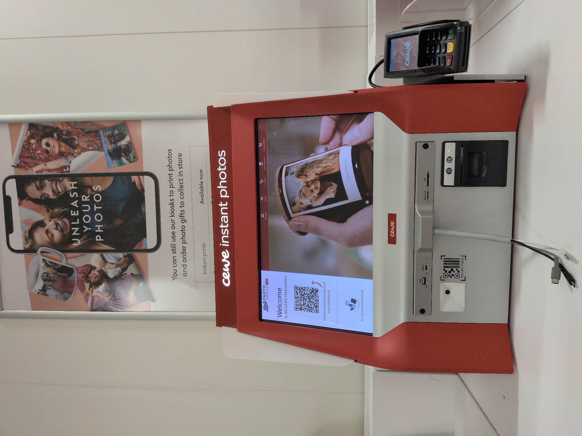 This is a medium shot of a CEWE photo kiosk, likely located in a Boots store in Telford, United Kingdom, given the branding on the kiosk and the presence of the "Boots PHOTO" logo. The kiosk is red and white and features a large touchscreen display. The display shows a welcome message and a QR code for transferring photos from a smartphone. Below the screen, there are various slots for memory cards, USB drives, and a print output tray. A card reader is attached to the side of the kiosk. Above the kiosk, a poster advertises "Unleash Your Photos" with various examples of photo products. The overall environment appears to be a retail setting with plain white walls. There are no people visible in the image.