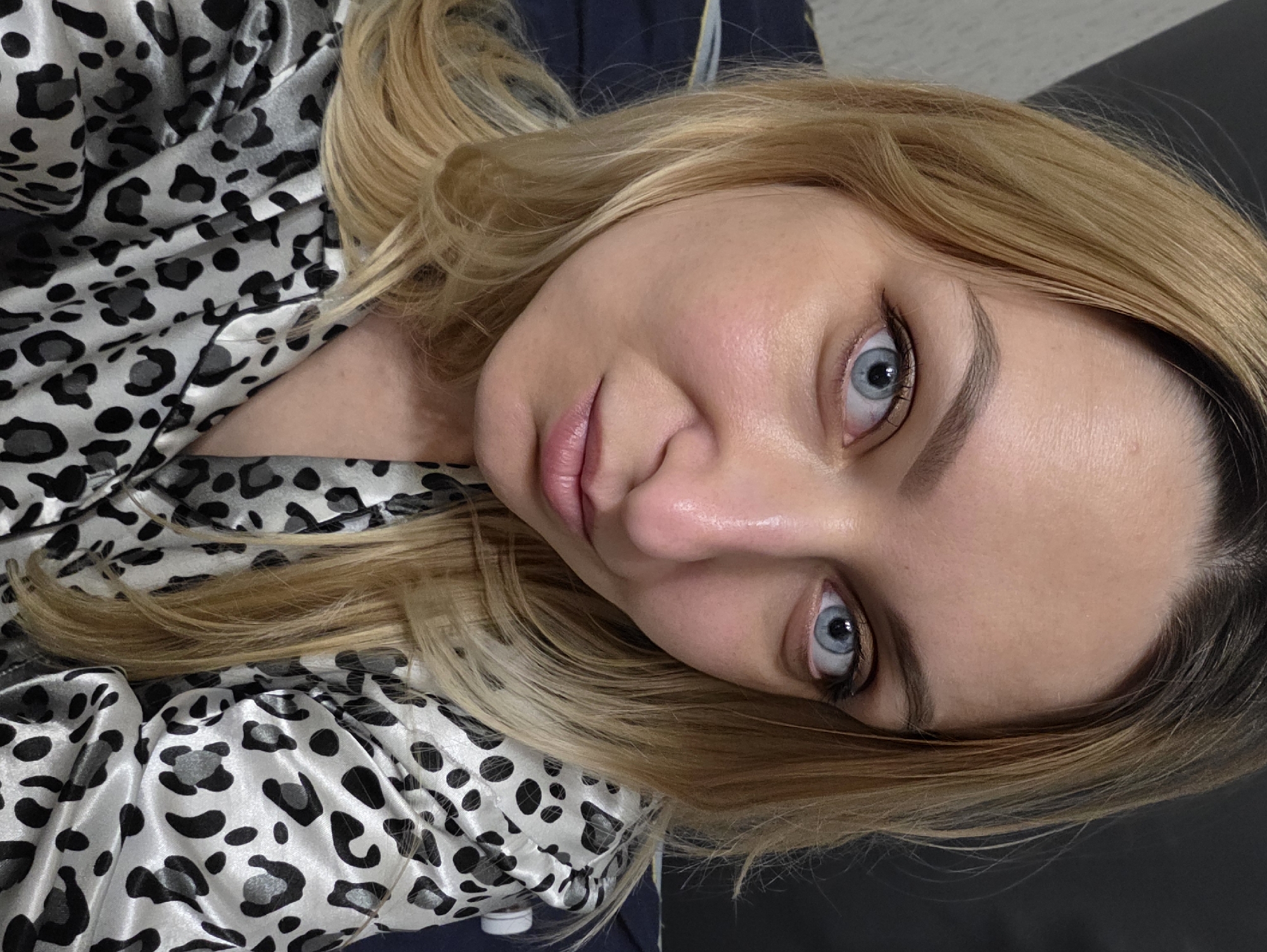 An adult female with blonde hair and striking blue eyes is depicted in a close-up, chest-up portrait. She has fair skin, a straight nose, and full, light pink lips that are slightly parted. Her gaze is directed centrally at the viewer. She is wearing a garment with a prominent black and grey leopard print pattern on a light, possibly satin-like, fabric. The background is dark and out of focus, providing no discernible details about the immediate surroundings. The image appears to be an indoor photograph, but no specific landmarks or environmental indicators are present to determine a precise location within Minsk, Belarus.