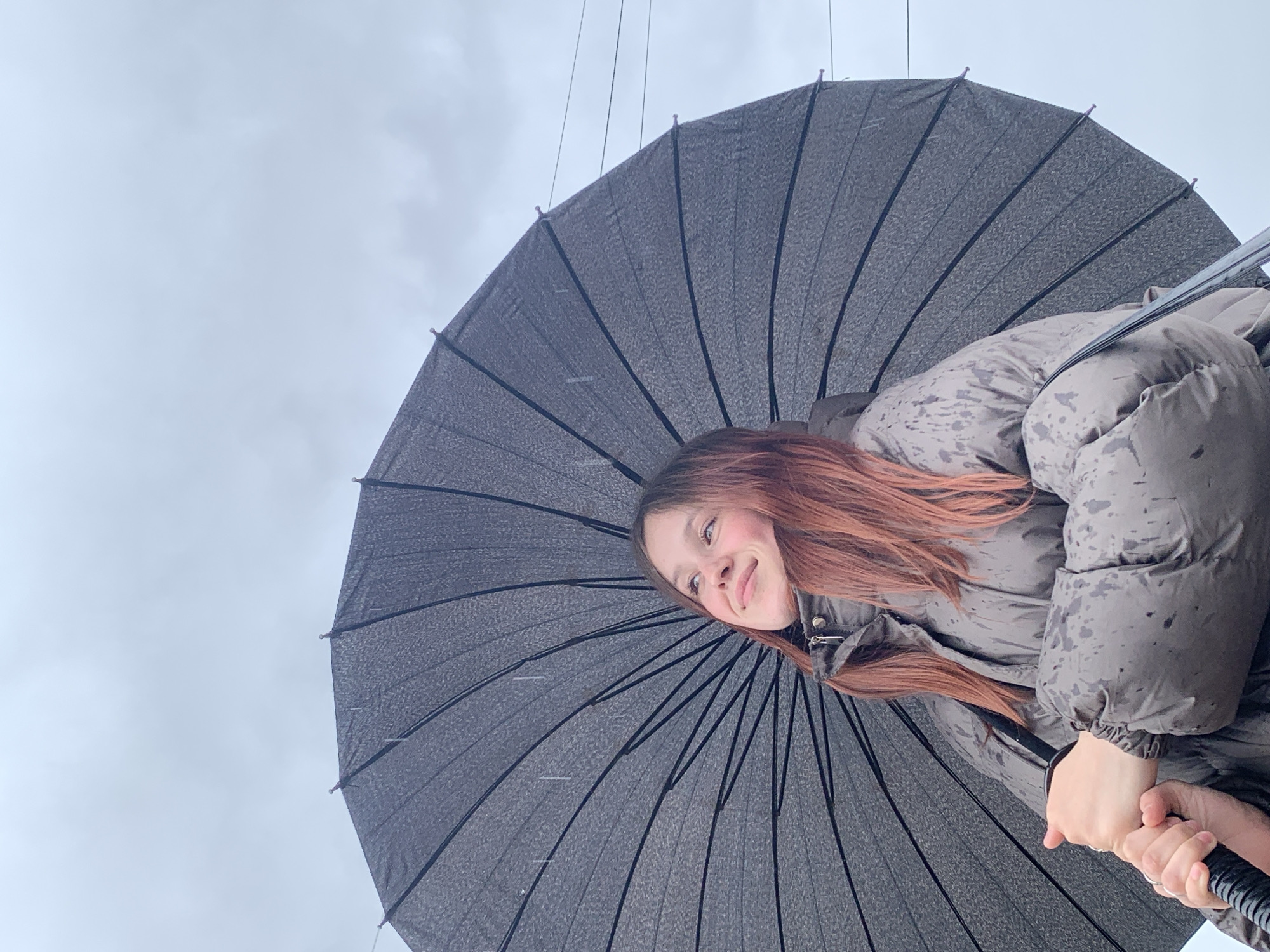A low-angle shot depicts a woman under a large, dark umbrella against a cloudy sky. The woman has fair skin and red-brown hair. She is wearing a gray puffer jacket. The umbrella, which is being held by the woman, has a textured surface and black ribs. The background is a pale gray sky. There is no indication in the image to suggest that it was taken in Фоки, Russia.