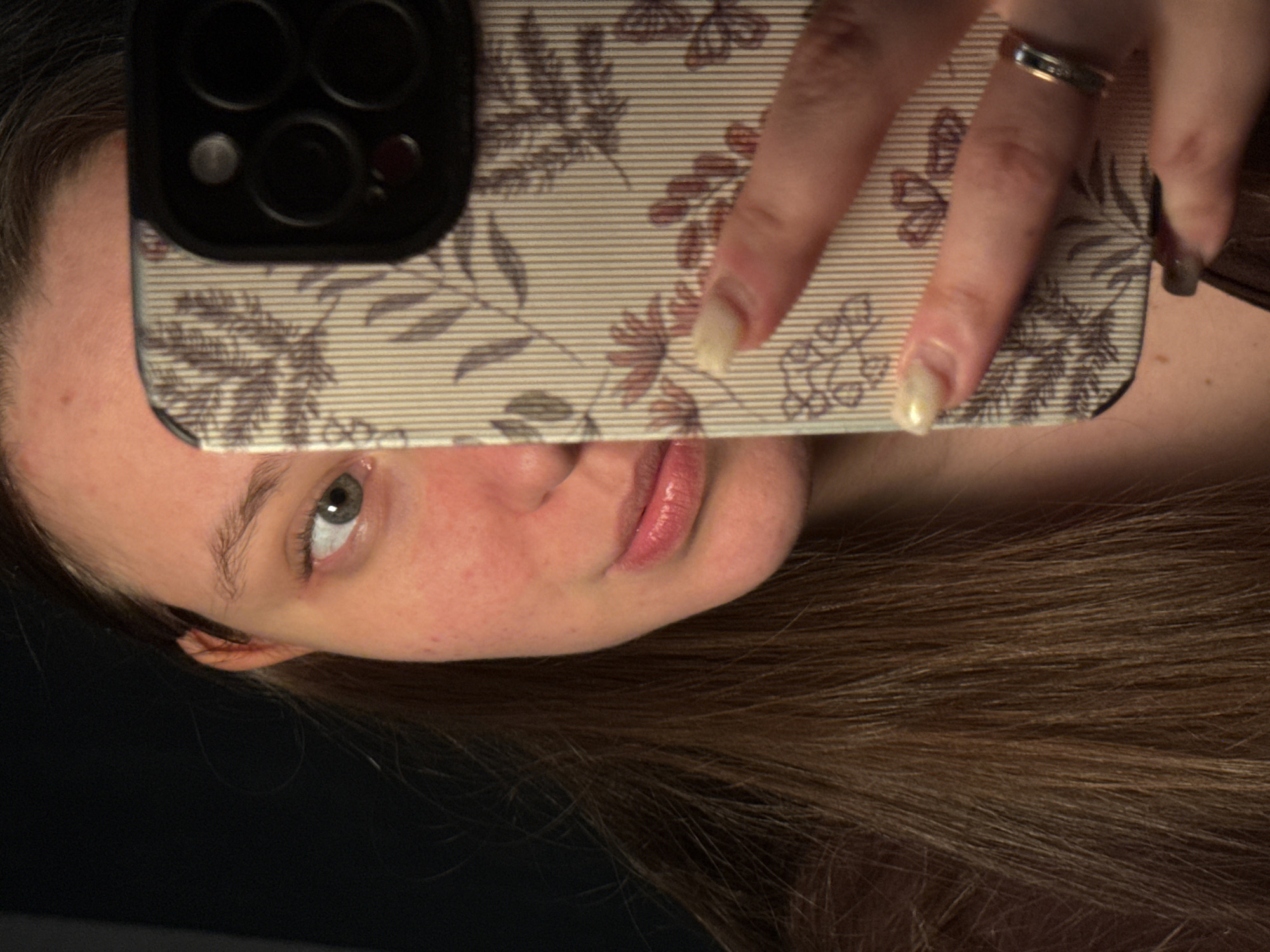 A person is taking a selfie in what appears to be a dimly lit indoor environment. The individual is holding a smartphone with a decorative case, partially obscuring their face. Their hair is brown, and their complexion is fair. A silver ring is visible on one of their fingers. Due to the limited visible context, providing the location within Krasnodar, Russia, is not possible.