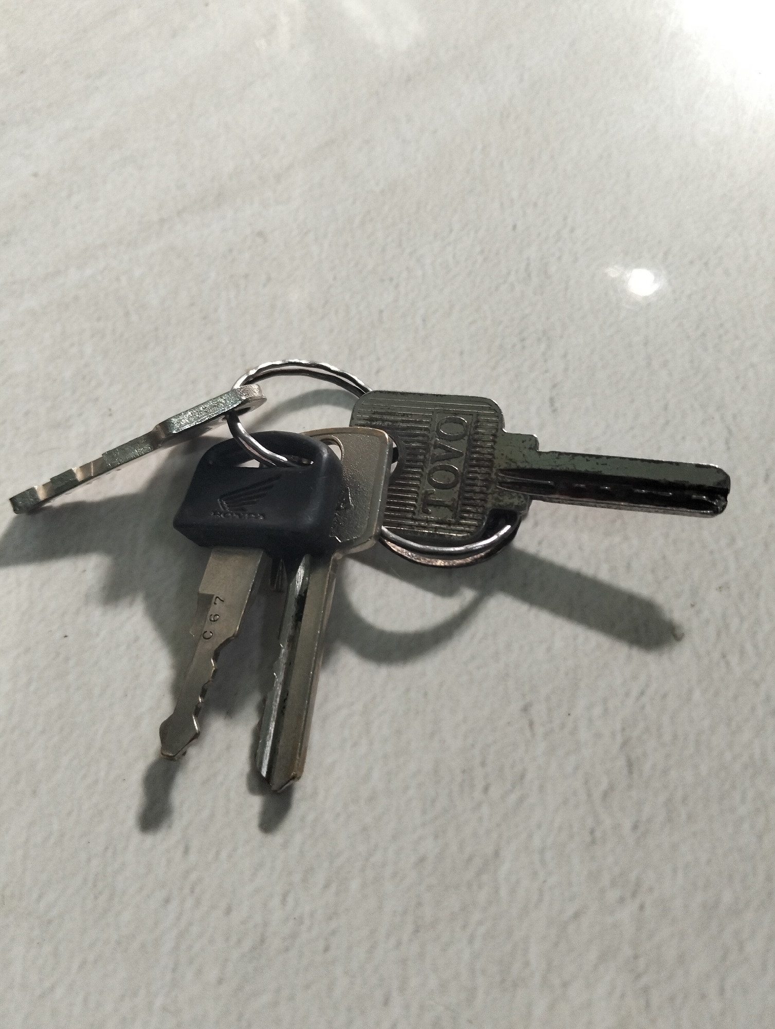 A close-up, overhead shot shows a keyring holding three keys, resting on a white, textured surface. One key has a black plastic head with the "HONDA" logo embossed on it, and the key blade displays the characters "C67". Another key has a silver head with "TOYO" printed vertically. The third key is entirely silver and has a notched blade. The surface appears to be a tiled wall or floor with a subtle grid pattern. Shadows are cast by the keys, indicating the direction of the light source. The image was taken in Paciran, Indonesia.