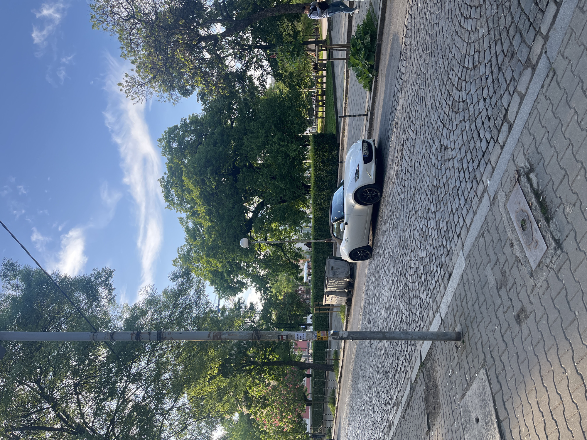 The image depicts a street scene in Sofia, Bulgaria. A white car is parked on a cobblestone street alongside a sidewalk. Trees and greenery line the street, with a glimpse of a park or open space visible beyond the foliage. A metal pole is positioned in the foreground, potentially a lamppost or traffic sign support. The sky is visible with light clouds. A person is standing on the sidewalk in the background. The location is likely a residential or mixed-use area, given the presence of trees, parked car, and sidewalk.