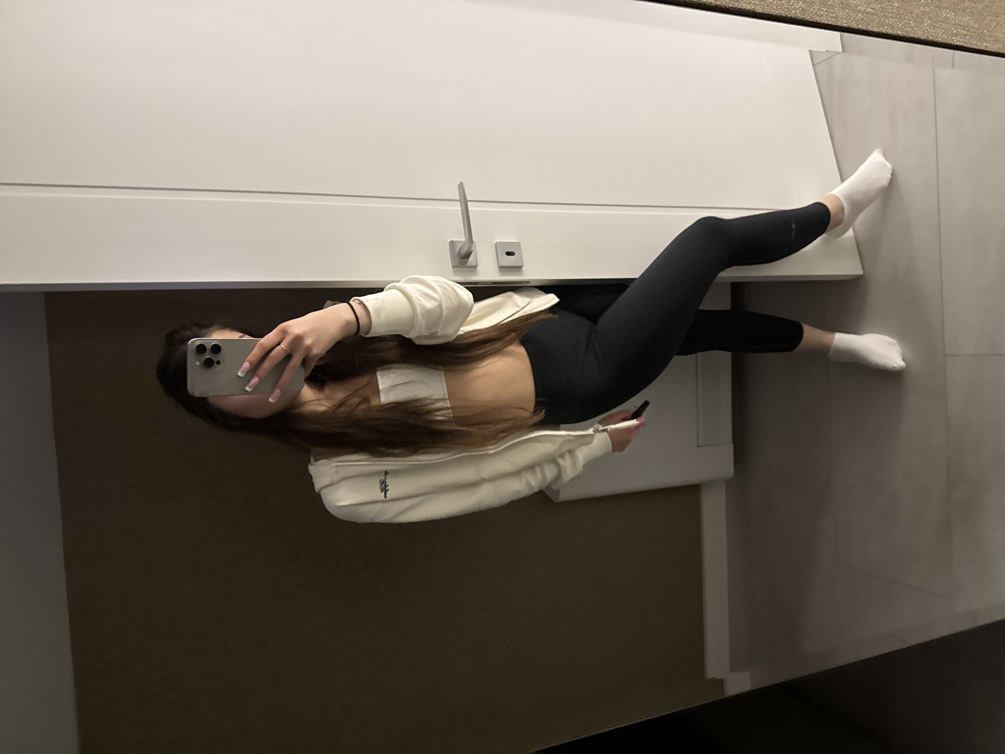 A young woman is taking a selfie in what appears to be a bathroom, possibly within a private residence. She is wearing black leggings, a white sports bra, a cream-colored zip-up jacket, and white socks. She is holding a smartphone with a gray case, partially obscuring her face. The background includes a white vanity or cabinet, a silver faucet, and gray floor tiles. The location within Plovdiv, Bulgaria, cannot be determined from the given image.