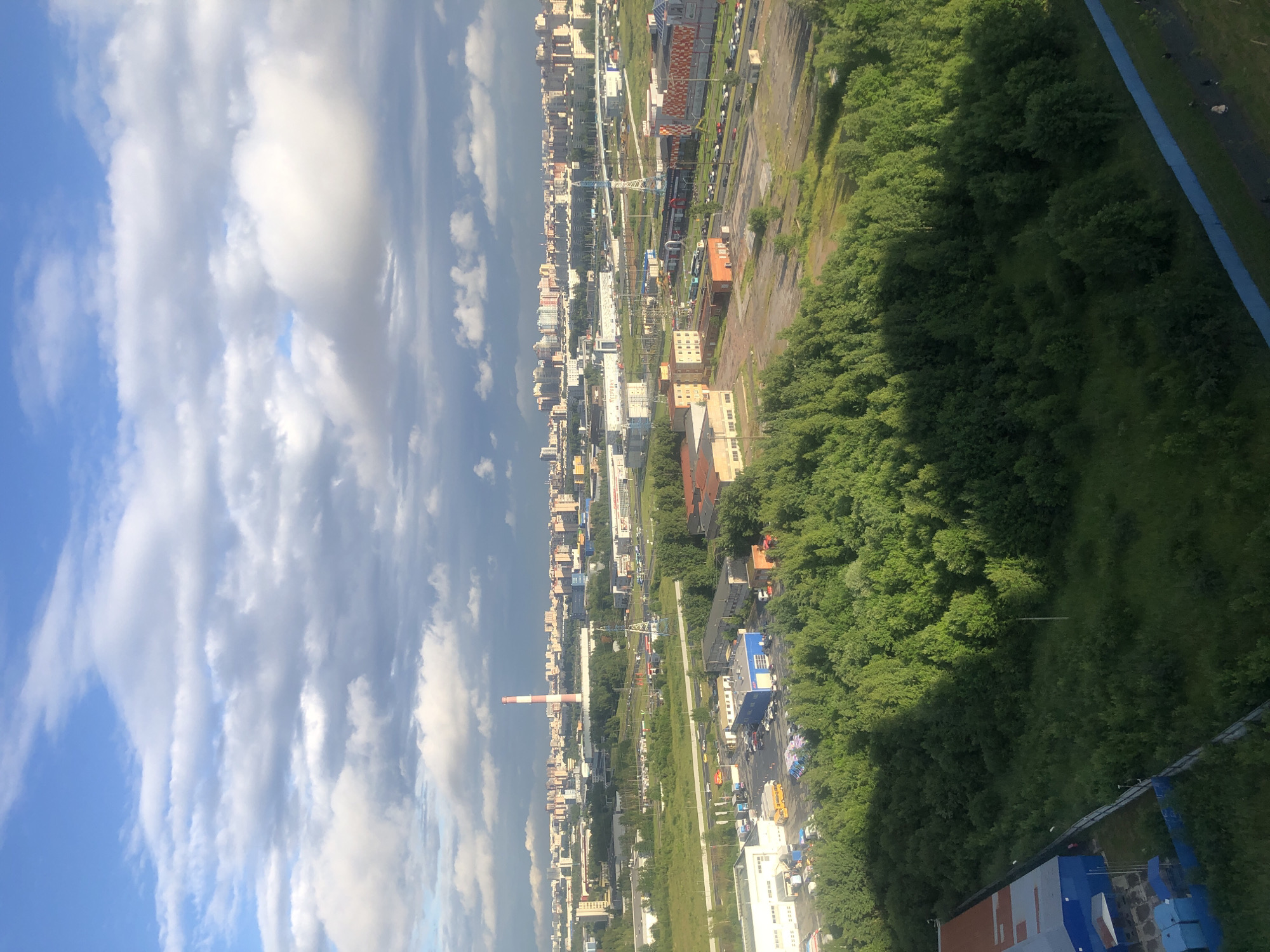 This is a high-angle, vertical shot of a city scene under a cloudy sky. The image is dominated by a mix of urban development and lush greenery.
On the left side of the frame, a vast expanse of sky is filled with dramatic, streaky clouds in shades of white and gray, against a bright blue backdrop. Below the sky, a sprawling city unfolds with numerous buildings of varying heights and architectural styles. Many appear to be residential apartment complexes, characterized by their multi-story designs and uniform windows. Interspersed among the buildings are hints of infrastructure, such as power lines and what looks like a tall industrial chimney with red and white bands. A construction crane is also visible within the urban landscape.
To the right of the city, a dense, vibrant green forest stretches along the edge of the urban area. The trees are full and leafy, suggesting a warm season. A road or pathway, marked by a bright blue line, runs alongside the forest, with a few vehicles visible on it. In the foreground, a blue and orange industrial-looking building with a sloped roof is partially visible.
The scene suggests a daytime setting, likely late morning or afternoon, given the brightness of the sky and the shadows cast by the trees. The weather appears to be partly cloudy, with the sun intermittently breaking through. There is no discernible activity or event taking place; it is a static view of the urban and natural environment. No text is legible in the image.