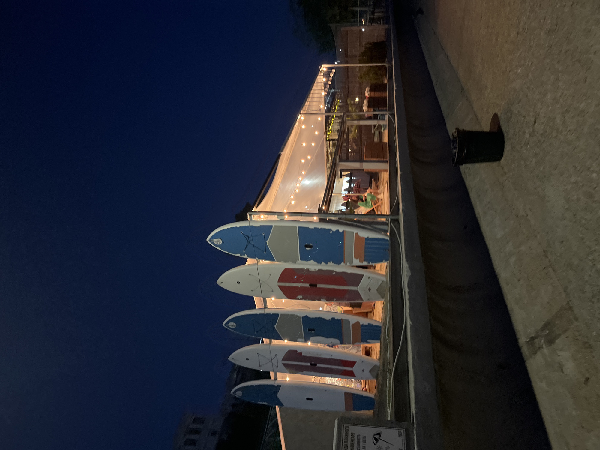 At dusk, on a concrete embankment in Sochi, Russia, a row of five paddleboards stands vertically. The paddleboards are painted with various patterns of white, blue, and red. Behind them, a covered outdoor seating area is illuminated by string lights, with a few people visible sitting at tables. To the left of the paddleboards, a sign with Russian text and an image of an umbrella is mounted on a post. A black trash can is situated on the embankment to the right of the seating area. The sky is a deep, dark blue, indicating it is nighttime.