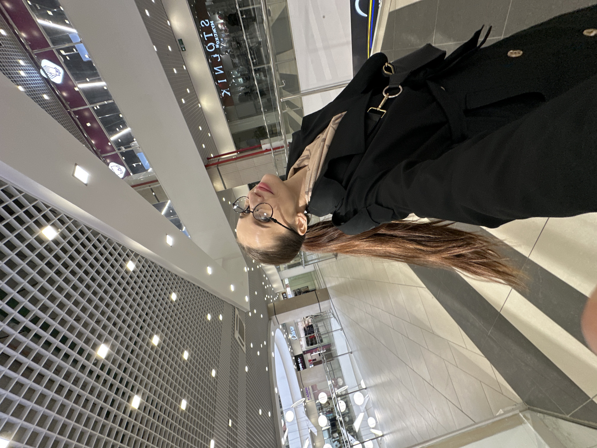 A young woman stands in what appears to be a modern shopping mall in Yekaterinburg, Russia. The image is taken from a low angle, looking slightly upwards, with the woman positioned on the right side of the frame, facing left in profile. She has long, straight brown hair pulled back into a ponytail that extends down her back. She wears clear, round-framed glasses and a black coat or trench coat over a lighter, possibly beige, patterned scarf or top. A black shoulder bag with gold-colored rings and clasps is visible on her right side.
The setting is an expansive indoor space characterized by contemporary architecture. The ceiling and upper walls feature a grid-like pattern with numerous small, square recessed lights, creating a bright, illuminated environment. Large, reflective panels or windows are visible above, some reflecting other parts of the mall and signage. The floor consists of light-colored tiles, some with a subtle geometric pattern, and distinct black and white striped sections. In the background, multiple levels of the mall are visible, with railings and various shop fronts.
There's no distinct activity occurring beyond the woman standing and looking upwards or to the left. The overall impression is one of casual observation or waiting within a bustling commercial space.
Notable details include the bright, artificial lighting suggesting an indoor daytime or well-lit evening setting. Several signs are reflected on the glossy surfaces of the mall's upper levels. Visible text reflections include "МАЖСКОЙ СТОЛИК" (MEN'S TABLE), "Charm", and "KAZAN" or "KAZAKH", likely referring to store names or brands. Some abstract white icons, resembling hands, are also reflected. The clean lines and reflective surfaces contribute to a sleek, modern aesthetic.