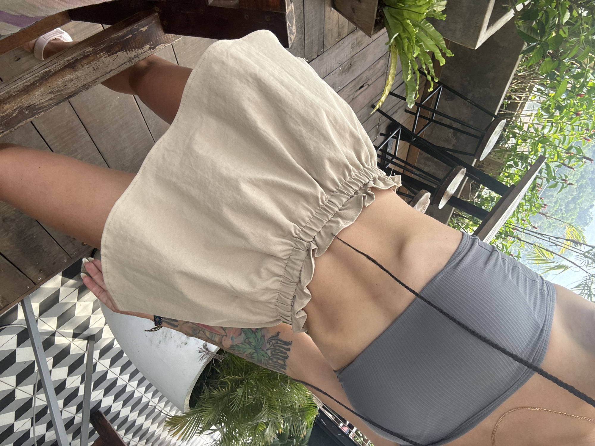 The image, captured from a first-person perspective, shows a person seated in an elevated outdoor setting, likely a cafe or balcony, with a view of a lush green mountainous landscape characteristic of Hà Giang 1 Ward, Vietnam. The person's legs, torso, and a tattooed arm are visible. They are wearing light beige shorts or a skirt with a ruffled hem and a grey ribbed bandeau top. A gold chain necklace and dark braided strings are also part of the attire. One foot, clad in a white sandal with a visible brand logo (likely Adidas), rests on a distressed wooden structure.
The immediate surroundings feature a wooden deck or floor. Adjacent to the person's feet, a distinct black and white geometric tiled floor, designed with a 3D cube pattern, is visible. The scene includes black metal chairs with wooden seats, and various green potted plants, such as ferns, contributing to a natural atmosphere. Bright, natural light suggests it is daytime with clear weather. The overall impression is one of relaxation amidst scenic surroundings.