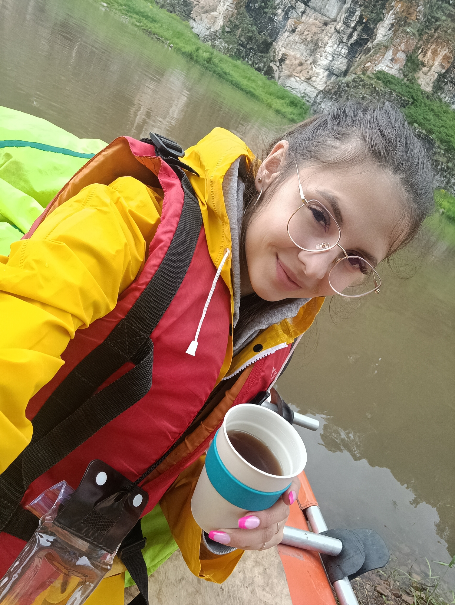A young woman with long, dark hair pulled back in a bun is taking a selfie while sitting in what appears to be a boat on a river. She is wearing a bright yellow jacket over a red and black life vest. She has on gold-rimmed glasses and is smiling at the camera. In her right hand, she holds a white mug with a teal band, and her fingernails are painted with pink and white French tips. A clear plastic pouch is visible on her left side, attached to the life vest. The background shows a body of murky water, with a steep, rocky cliff face covered in green vegetation. The location is identified as Idrisovo, Russia.