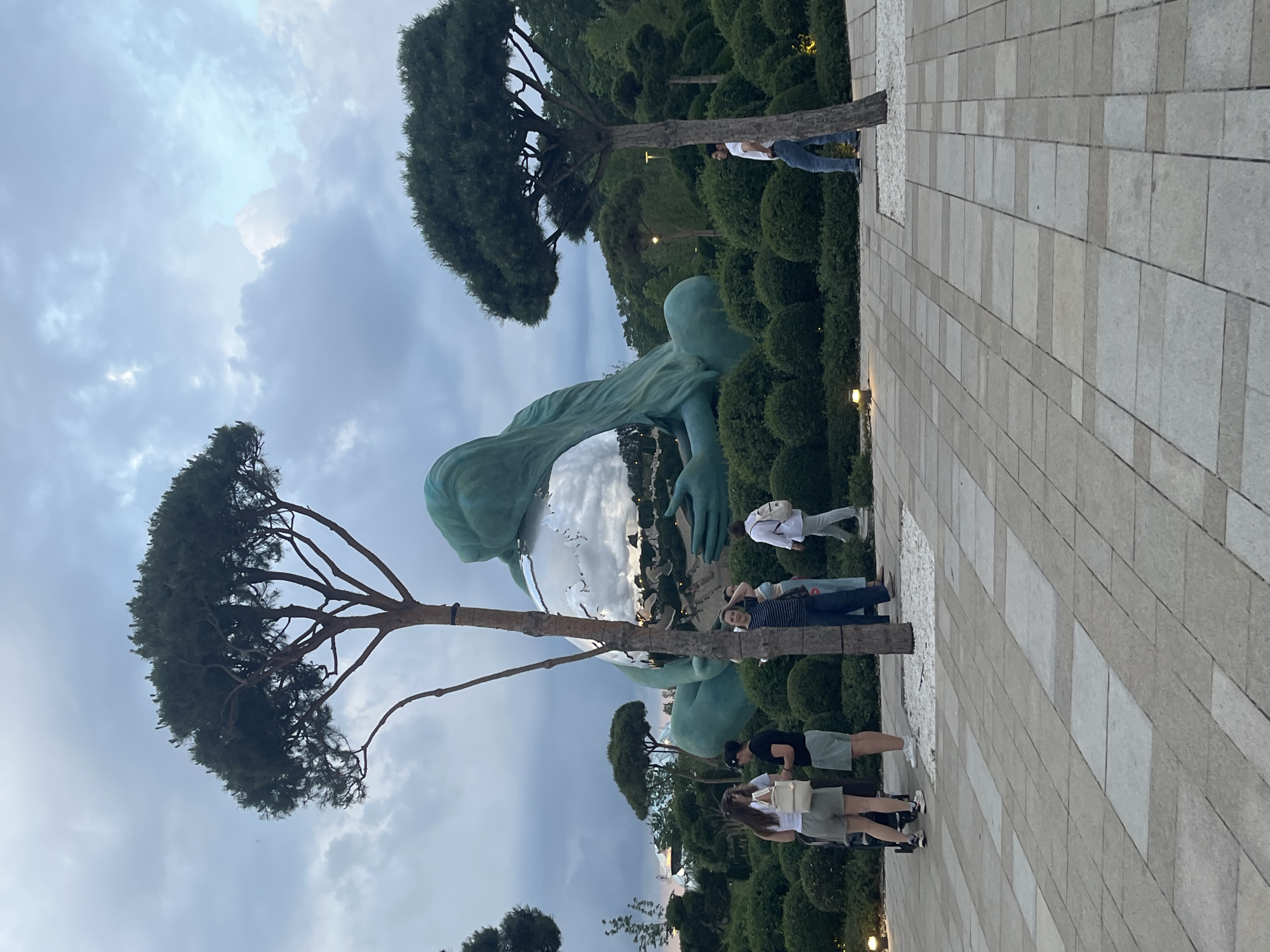 This is a full-shot, eye-level, outdoor photograph taken in the city of Krasnodar, Russia. The image captures a large, artistic sculpture of a woman cradling a reflective sphere in her hands, set against a backdrop of lush greenery and a cloudy sky. The sculpture is a dominant feature, with its turquoise hue and polished surface reflecting the surroundings. In the foreground, a paved walkway made of rectangular stone tiles is visible, with a few people scattered throughout the scene.
On the left side of the walkway, a group of four people is gathered, appearing to be in conversation. Two women in casual attire, one with a white purse, stand facing a person wearing a black t-shirt and shorts. Further down, a child in a stroller is partially visible.
In the middle ground, to the right of the sculpture, a lone man in a white t-shirt and blue jeans stands near a tree, looking towards the sculpture. Another individual, partially obscured by a tree, appears to be taking a photograph.
The environment is characterized by manicured bushes and trees, with soft lighting from strategically placed outdoor lamps. The overall atmosphere suggests a public park or a similar recreational space designed to showcase art and nature. The sky is overcast, with variations in cloud density, indicating a diffused natural light. The "Golden Domes" of the Cathedral of the Nativity of the Blessed Virgin Mary, a prominent landmark in Krasnodar, can be seen in the distant background.