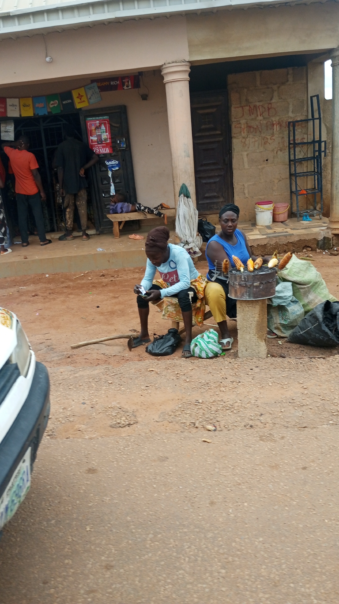 The image depicts an outdoor scene in Reifild-Ist, Nigeria, likely a street or market area. Two women are the central focus. The woman on the left, wearing a light blue long-sleeved shirt and dark pants, is seated on a stool, looking down at a mobile phone in her hands. She has a brown head covering. Next to her sits a black bag and a hoe-like tool on the ground. To her right, another woman, wearing a blue sleeveless top and yellow pants, is seated, tending to a charcoal grill laden with corn on the cob. She has a black head covering. Behind the women, a rustic structure with a weathered brick wall and wooden doors is visible. In front of this structure, several people are standing or sitting. One person in an orange t-shirt and dark pants stands near a shop entrance, which displays various colorful posters and products. Another person in dark clothing is seen behind them. A child is lying on a wooden bench in front of the shop. Bags of what appear to be produce or goods are stacked near the grill and the shop. The ground is composed of dirt and gravel, with some areas of cracked concrete. A portion of a white vehicle is visible in the lower left corner of the frame. The overall atmosphere suggests a typical day in a Nigerian urban or semi-urban setting with commercial activity.