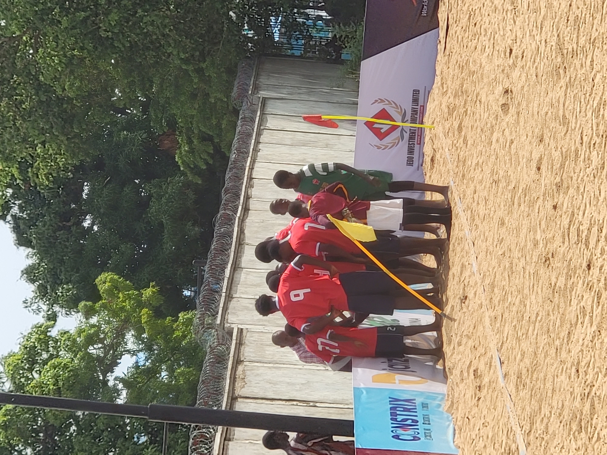 A vertical shot shows a group of people on sand in Unguwar Sarki, Nigeria. The team closest to the camera are wearing red uniforms. The team closest to the wall are wearing green uniforms. Behind the teams are banners for JEDO Investment Company Limited, CONSTRIX, and JOIZ. A tall wall with barbed wire on top is near the players. Trees are next to the wall. A pole is on the left.