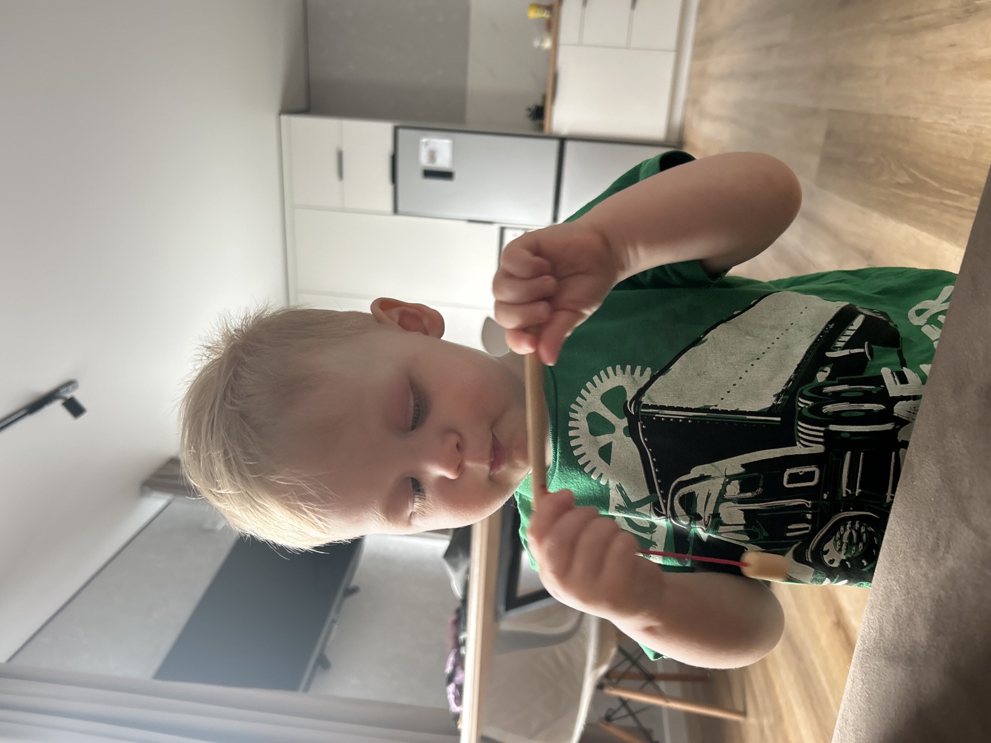 A young blonde-haired child is shown in a focused, candid moment indoors, likely in a combined kitchen and living space in Chaikovsky, Russia. The child, appearing to be a toddler or preschooler, is wearing a vibrant green t-shirt featuring a detailed graphic print of a truck or large vehicle, including a prominent spoked wheel. Their gaze is directed downwards at a thin, light-colored stick or skewer they are holding, possibly engaged in eating a snack or playing.
The background reveals a modern and well-lit home interior. On the right side of the frame, a contemporary kitchen area is visible with sleek white cabinets, both upper and lower, and a silver-toned refrigerator. The flooring throughout is a light wooden laminate or plank. To the left, elements of a living area can be seen, including part of a television screen and a black ceiling-mounted light fixture. A neutral-toned sofa or cushioned surface is partially visible in the lower right foreground. The overall impression is one of quiet concentration during a daytime activity. No legible text is visible in the image.