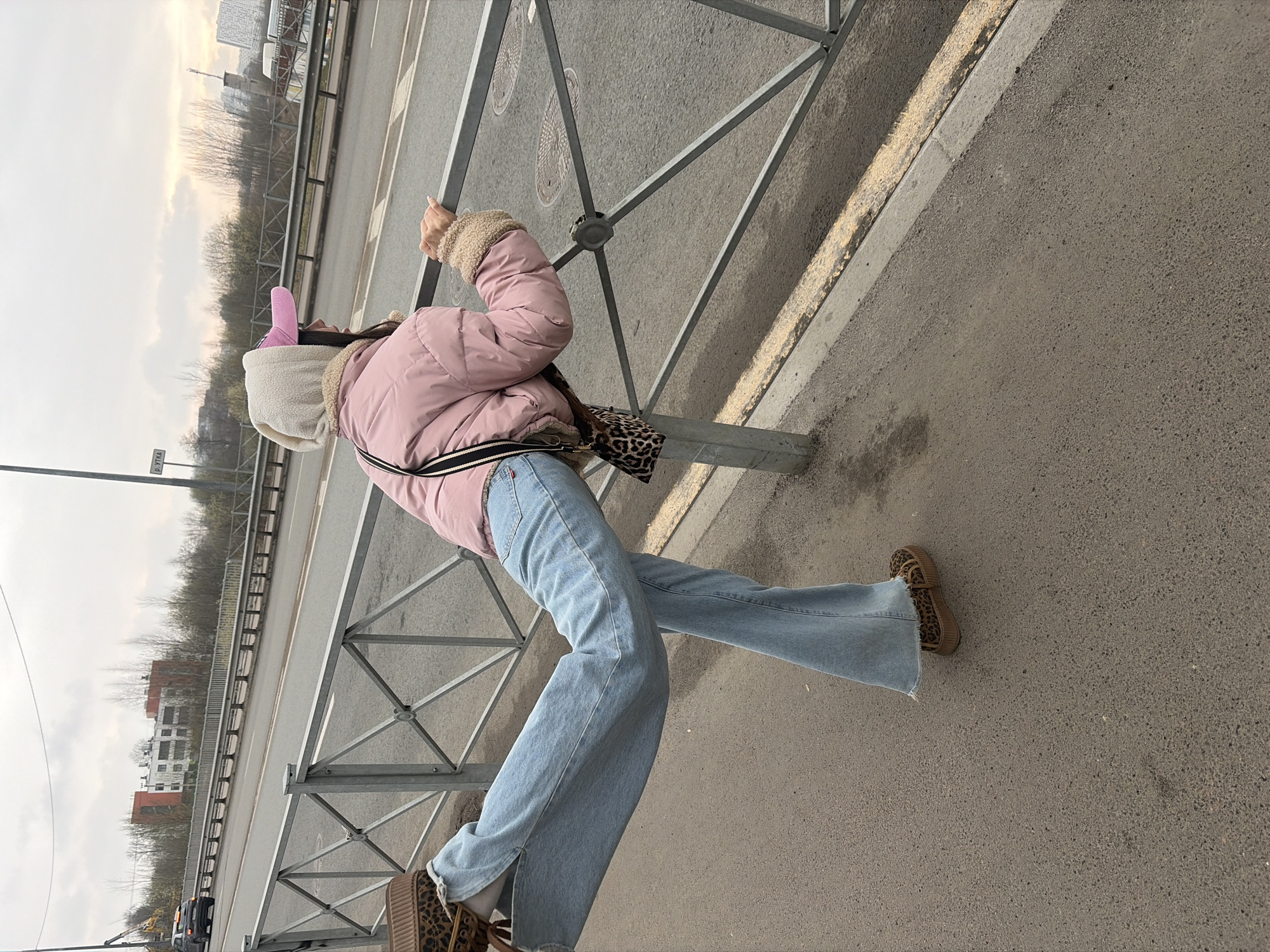 A person is standing near a metal guardrail in what appears to be an urban environment. The individual is wearing a pink puffer jacket with fur trim around the cuffs, light blue jeans, and leopard-print shoes. They also have on a white hat with pink bunny ears. A leopard-print bag is slung over their shoulder. The background shows a road and buildings. Based on the roadside signs and architecture, the location might be Saint Petersburg, Russia. The person seems to be leaning on the guardrail, facing away from the camera.