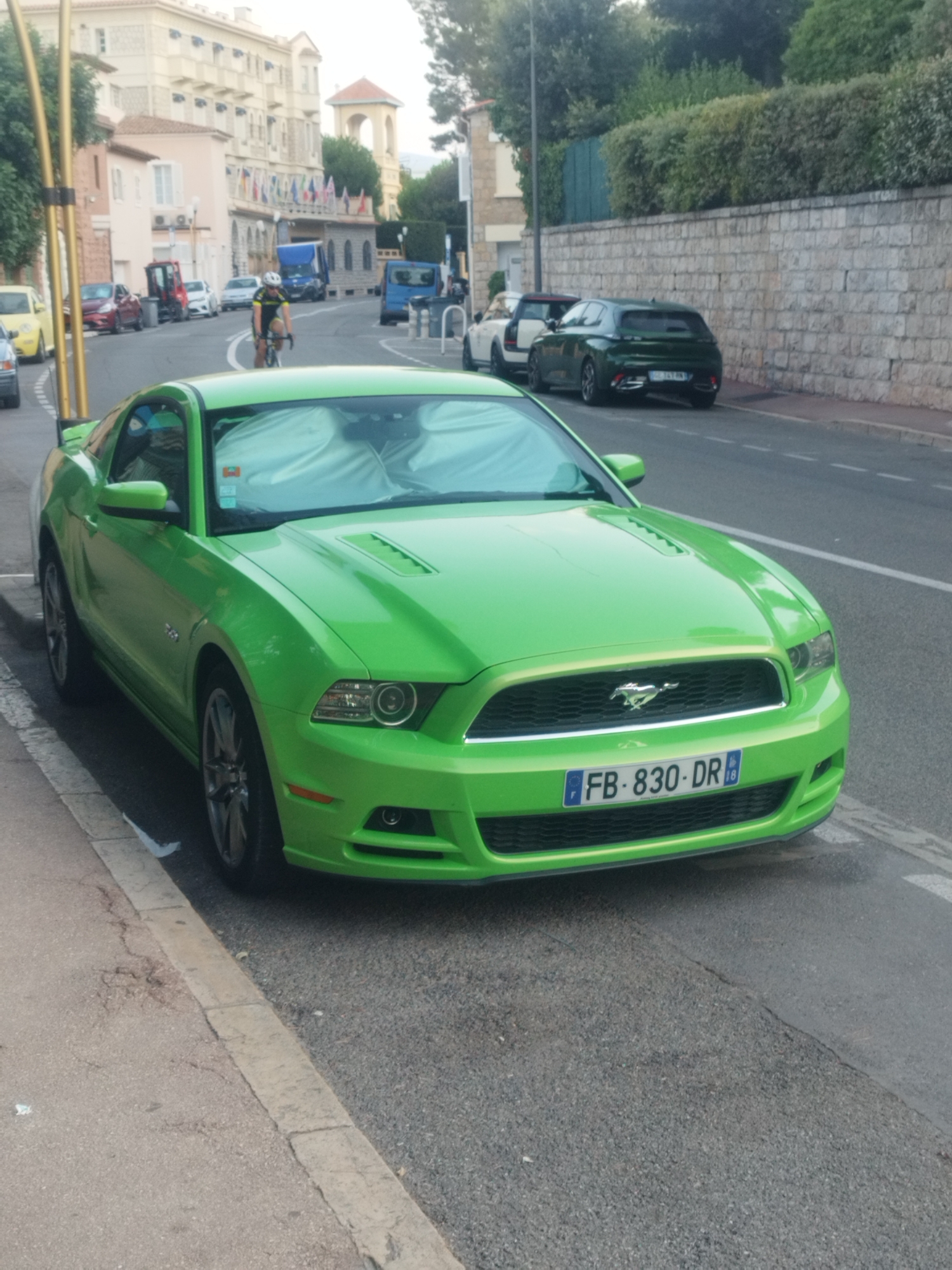 The image shows a bright green Ford Mustang parked on the side of a street in Antibes, France. The car is facing forward, with its front bumper and grille clearly visible. The license plate reads "FB-830-DR". In the background, there are several buildings with flags, suggesting a public area or event. A cyclist is also visible in the middle ground, riding on the street. The street appears to be paved, with a sidewalk on the left. The overall scene suggests a sunny day in a European city.