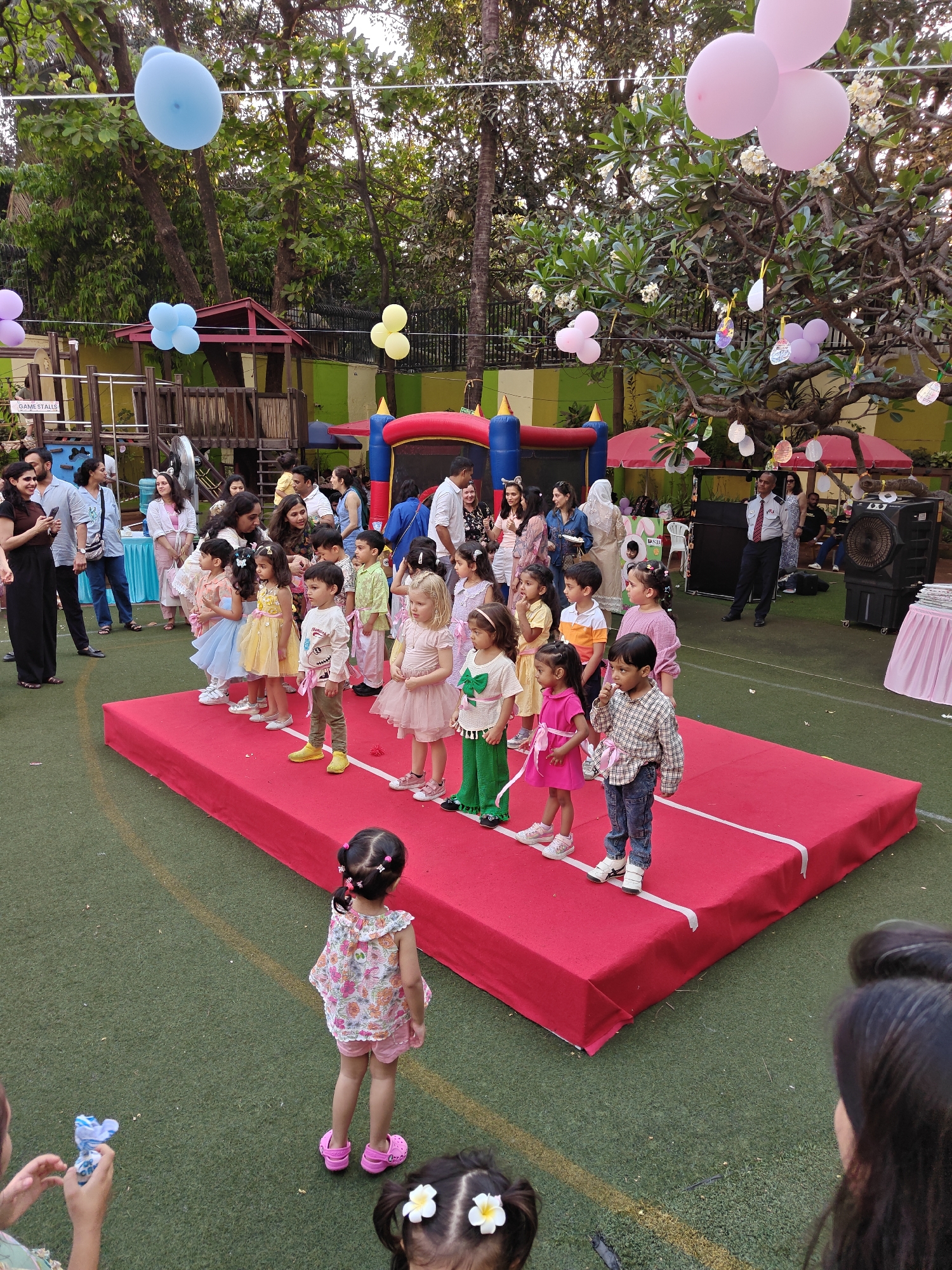 An outdoor children's event is depicted on a green artificial turf area, identified by a partially visible "Billabong High International School" sign in the background, indicating a location in Mumbai, India.
In the foreground, a raised, rectangular red platform, approximately 2.5 meters wide and 5 meters long, serves as a stage. White tape marks the perimeter of the platform. A line of approximately 12-15 young children, aged roughly 3-6 years, stand on this platform, facing generally towards the right side of the frame. The children are dressed in various festive outfits, including dresses with full skirts, patterned shirts, and colorful shoes. To the immediate left of the stage, an adult appears to be interacting with one of the children. Directly in front of the stage, closer to the viewer, a young girl with dark hair tied in pigtails, wearing a floral top and pink shorts with pink Crocs, stands with her back to the camera, observing the stage. Partially visible in the bottom left is another child's hand holding a crinkled blue object, and in the bottom right, the top of another child's head with white flower hair clips.
The midground features a crowd of approximately 30-40 adults and older children standing on the green turf, observing the event. Many adults are holding mobile phones, seemingly recording or photographing the children on stage. On the far left, a woman in a black top and wide black pants stands, looking at her phone. Behind the crowd, various structures are visible: a blue, red, and yellow inflatable bouncy castle, a wooden playground structure with a "GAME STALLS" sign, and multiple red canopies with tables underneath. A security guard in a light blue shirt, dark tie, and dark pants stands to the right of the canopies. A large black portable cooler/fan unit is positioned near the security guard, and a table draped with a pink tablecloth is partially visible on the far right.
The background consists of a lush green environment dominated by large trees with dense foliage, some bearing white flowers. Numerous blue, pink, yellow, and light purple balloons are tethered and float above the scene, some individually and others in bunches. Decorative paper cutouts or lanterns hang from the branches of one tree. A brightly painted wall, green at the top and yellow below, forms the boundary of the property behind the trees and structures. The ground throughout the visible area is green artificial turf with a yellow line visible on the left.