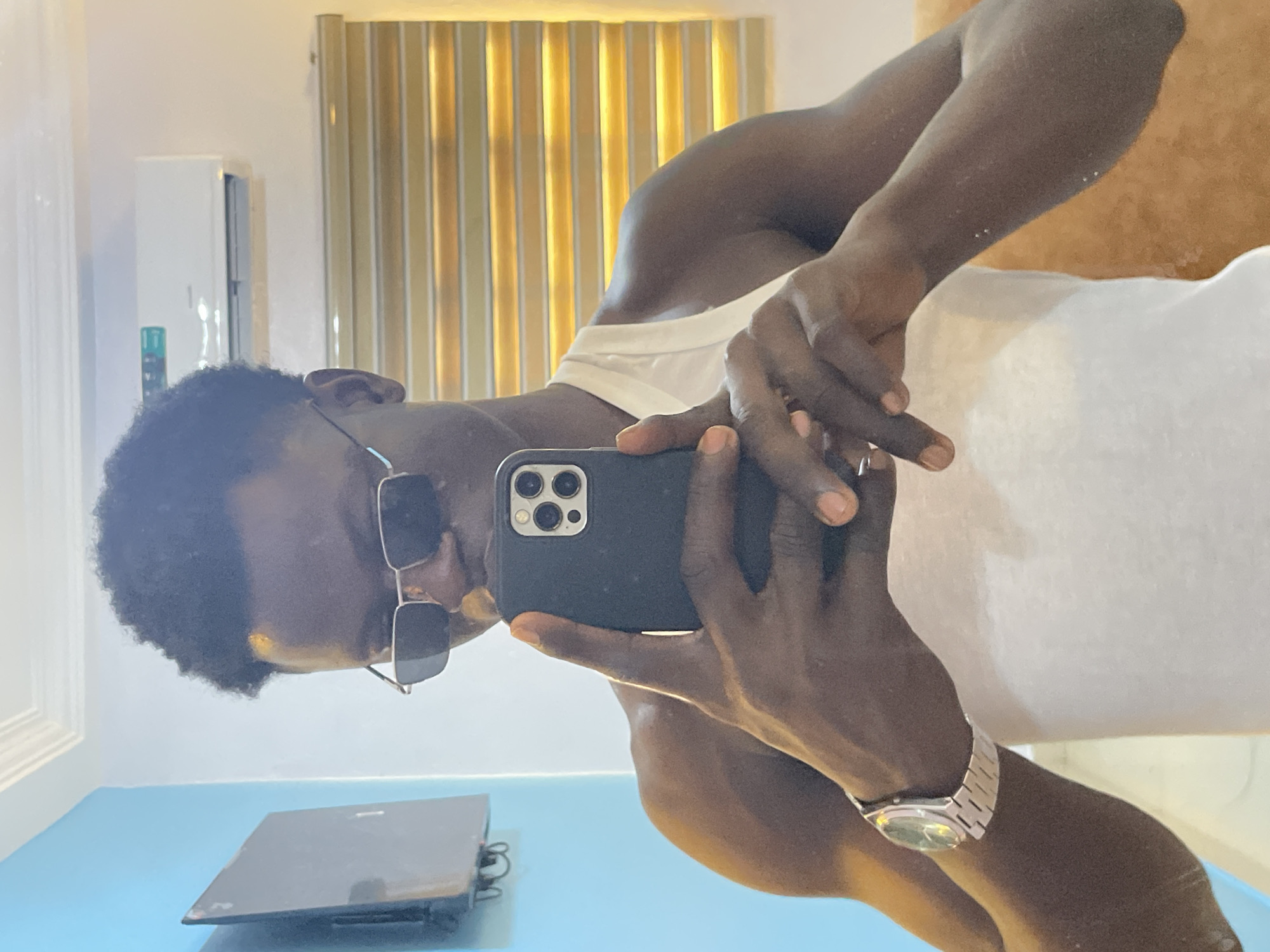 A selfie is being taken by a dark-skinned male, wearing a white tank top and sunglasses. He has a silver wristwatch on his left wrist. The background reveals a room with vertical striped wall panels and a blue table surface with a laptop. Based on the request, this image is presumed to be taken in Badagry, Nigeria.