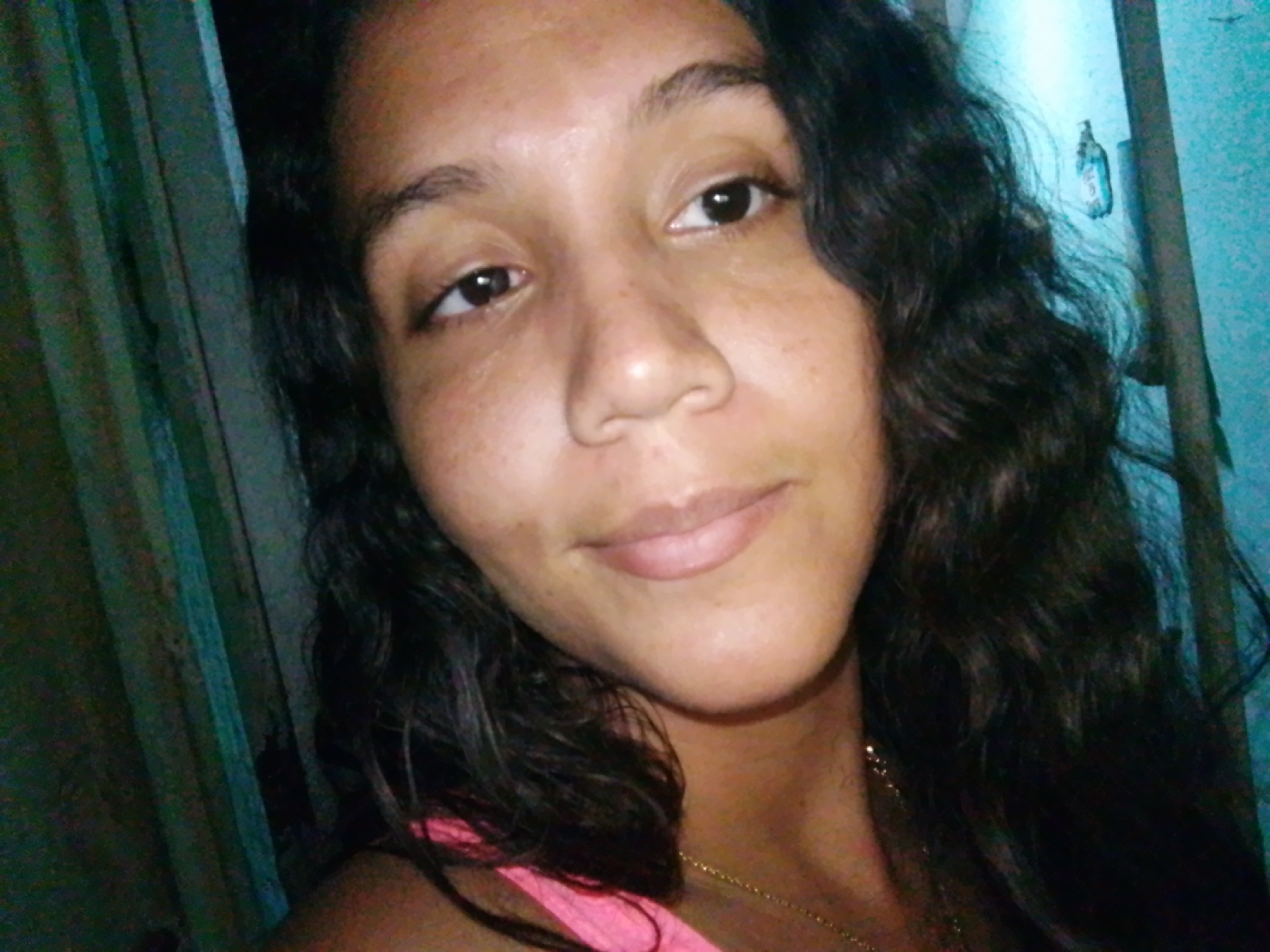 A close-up, head-and-shoulders shot of a woman with dark curly hair, dark eyes, and tan skin. She has a slight smile and is wearing a pink top and a gold necklace. The background appears to be an interior space with teal walls and a curtain. The image lacks sufficient detail to confidently place the scene within Juan Griego, Venezuela.