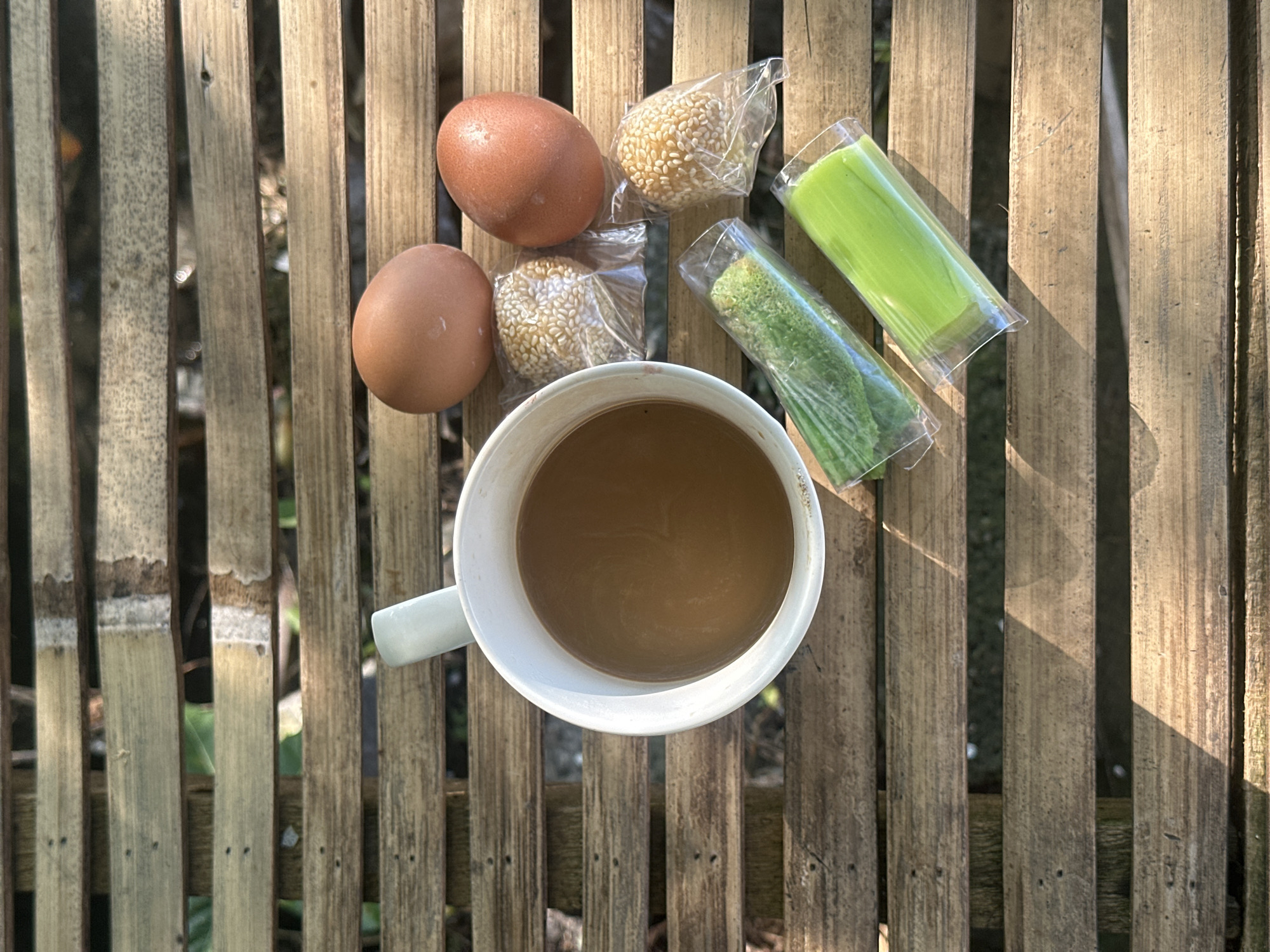 A high-angle shot reveals a breakfast scene set on a bamboo slat surface, likely a table or mat. Two brown eggs, two individually wrapped sesame balls, and two cylindrical green desserts encased in clear plastic are arranged near a white mug filled with a light brown beverage, likely coffee. The background is a blur of foliage, indicating an outdoor setting. The scene is illuminated by sunlight, creating distinct shadows on the bamboo surface. The precise location of the photograph is in North Kuta, Indonesia.