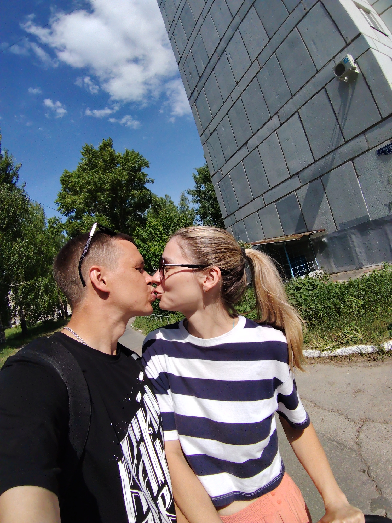 A couple is kissing in an outdoor setting. The male has short hair, sunglasses pushed back, and wears a black t-shirt with white patterns and a silver necklace. He also has a black backpack. The female has blonde hair in a ponytail, glasses, and a striped white and blue t-shirt. She's wearing peach-colored shorts. The background includes a large grey building with a surveillance camera and a small window with bars. Green trees and a blue sky with scattered clouds are visible above. The couple stands on a cracked asphalt surface. The building at the right of the image has an address plate which reads “ЁЗОВА 5/31”. Based on this address, the location is in the city of Chaikovsky, Perm Krai, Russia.
