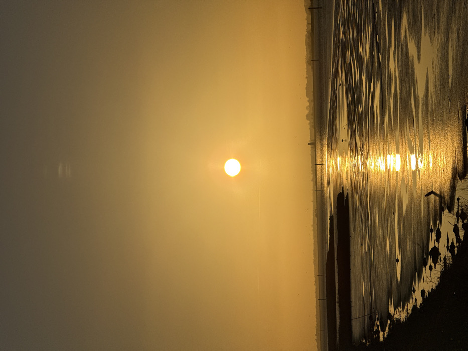 The image depicts a static landscape view of Breydon Water, Great Yarmouth, United Kingdom, during either sunrise or sunset. A brilliant, distinct white-yellow sun disc is positioned slightly right of the frame's vertical center, approximately two-fifths from the top edge. This sun casts a prominent, shimmering golden vertical reflection directly across the water's surface, extending towards the bottom-center of the image.
The sky, occupying the upper two-thirds of the frame, exhibits a gradient of golden-orange coloration directly around the sun, transitioning to a slightly darker, muted orange-brown towards the top and left edges. Faint, wispy atmospheric streaks or contrails are discernible in the upper-left quadrant of the sky.
The lower third of the image features the tidal estuary. The immediate foreground on the left consists of a dark, indistinct bank or shoreline with minor undulations and sparse, dark vegetation. The dominant feature of the lower half is the expansive, textured mudflats and shallow water of Breydon Water, largely exposed due to low tide. These surfaces display intricate patterns and reflect the golden light. Several thin, dark, vertical marker poles are sparsely distributed across the water in the mid-ground.
On the distant horizon, a low-lying, dark silhouette of land or distant infrastructure is visible. A prominent, dark, rounded industrial structure, consistent with a silo or tower, is clearly silhouetted on this distant horizon, positioned to the right of the sun's reflection. Multiple small, dark specks, identifiable as birds, are scattered across the mudflats and in the distant sky. No human figures or dynamic actions are observable.
