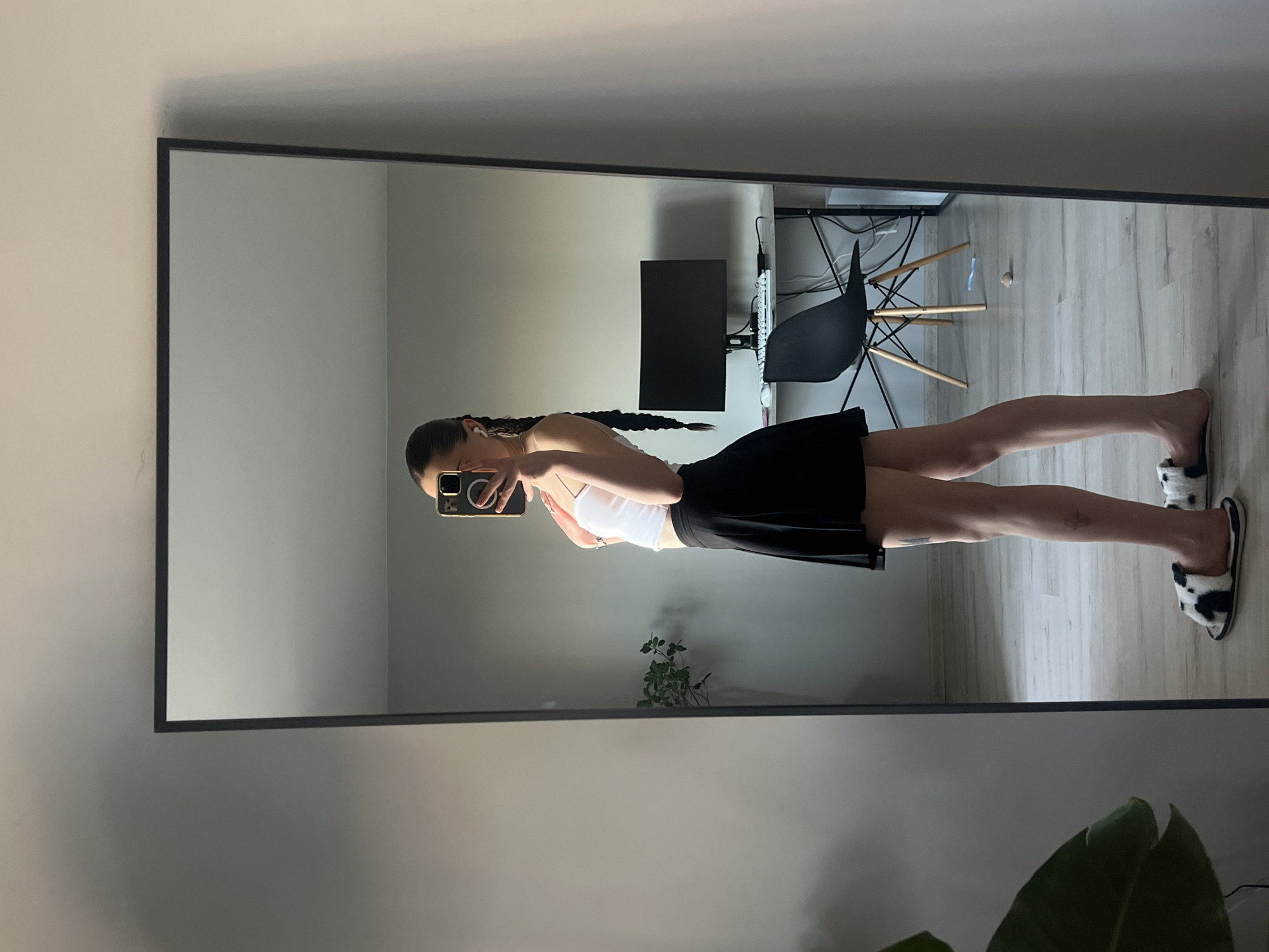 A young woman stands in front of a large mirror, taking a selfie. She has long dark hair braided down her back. She is wearing a white tank top and a black skirt. Her right foot is visible, wearing a fuzzy black and white slipper. Her left foot is also visible, wearing a similar slipper. The mirror reflects a room with light-colored walls and a light-colored wooden floor. A desk with a computer monitor and a black chair are visible in the reflection. A plant is also visible in the reflection, to the left of the woman. The lighting suggests it is daytime. The woman appears to be casually dressed in her home. There is no visible text in the image.