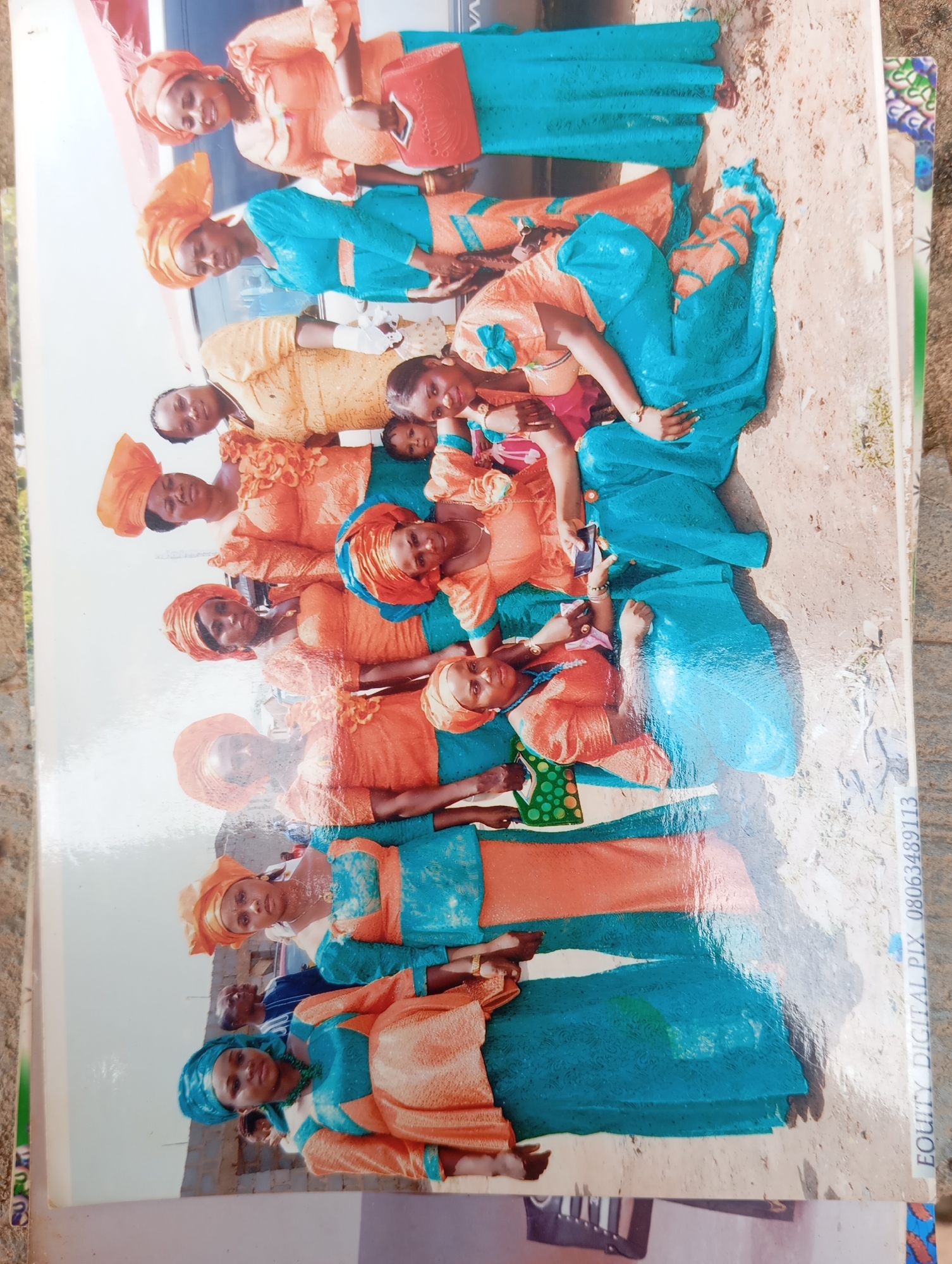 The image shows a group of women and a child in Karsana 2, Nigeria. The women are dressed in matching traditional Nigerian attire, consisting of brightly colored orange and blue fabrics. Some women are wearing headwraps in coordinating colors. They are arranged in a layered formation, suggesting a posed photograph. The background appears to be outdoors, with dirt and sand on the ground, and some hints of greenery and possibly some brick structures. One woman in the foreground is holding a mobile phone. The overall scene suggests a gathering or celebration. The photograph is oriented vertically, and a watermark with "EQUITY DIGITAL PIX 08063489113" is visible along the right edge.