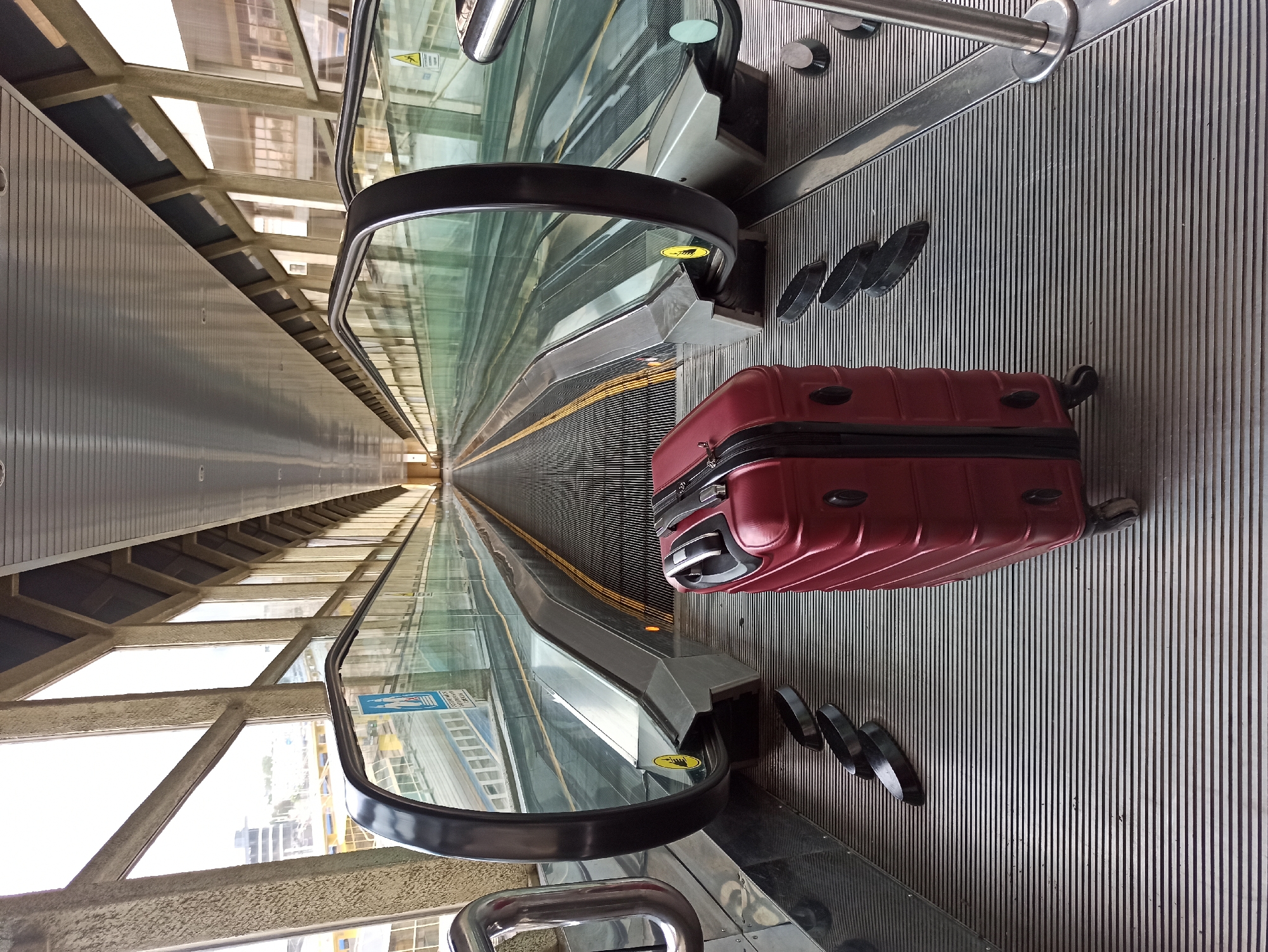 A red suitcase sits on a ribbed metal surface of a moving walkway at an airport. The walkway is enclosed by glass barriers and an overhead structure with angular supports. The walkway stretches into the distance, with similar moving walkways on either side. The overall environment appears to be an airport terminal, designed for efficient passenger transit. The scene is devoid of people. The image appears to be taken at eye level, looking forward along the walkway.