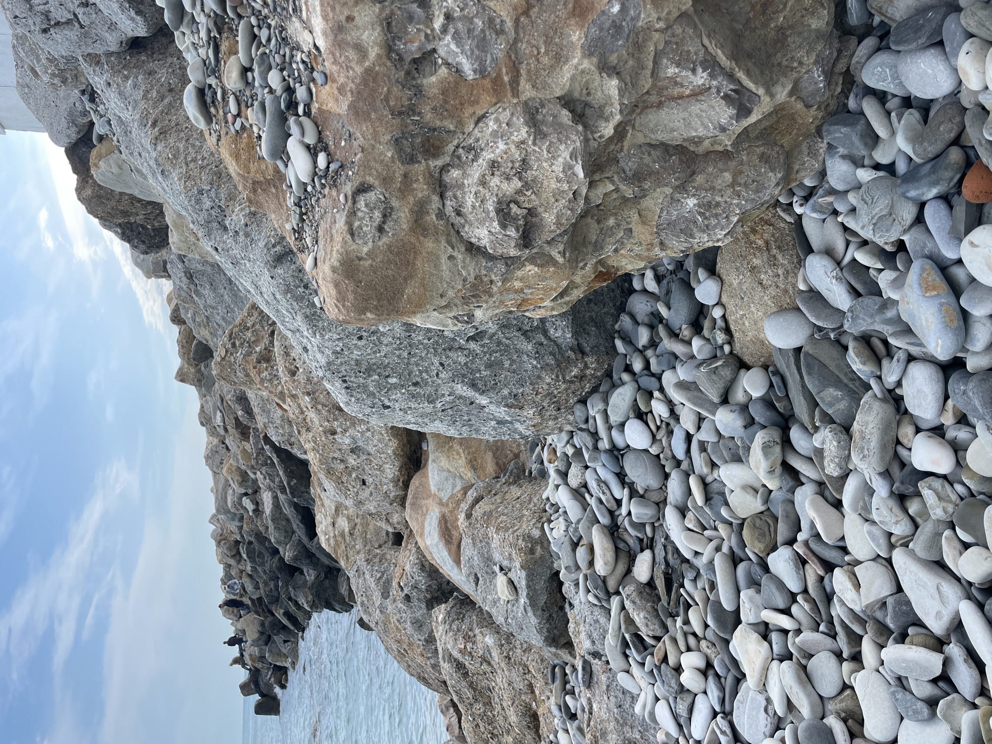 The image depicts a rocky shoreline along the Black Sea in Olginka, Russia. The foreground is dominated by a jumble of grey and brown rocks of various sizes, some embedded with smaller pebbles. In the middle of the frame, loose pebbles of grey and white cover the ground. At the lower left of the image, the rocks descend to meet the sea, which is visible as a light blue expanse under a light blue sky with scattered clouds. On the rocks jutting into the water at the bottom, several small figures, possibly people or birds, are visible, perched on the rocks. The photograph is rotated counterclockwise.
