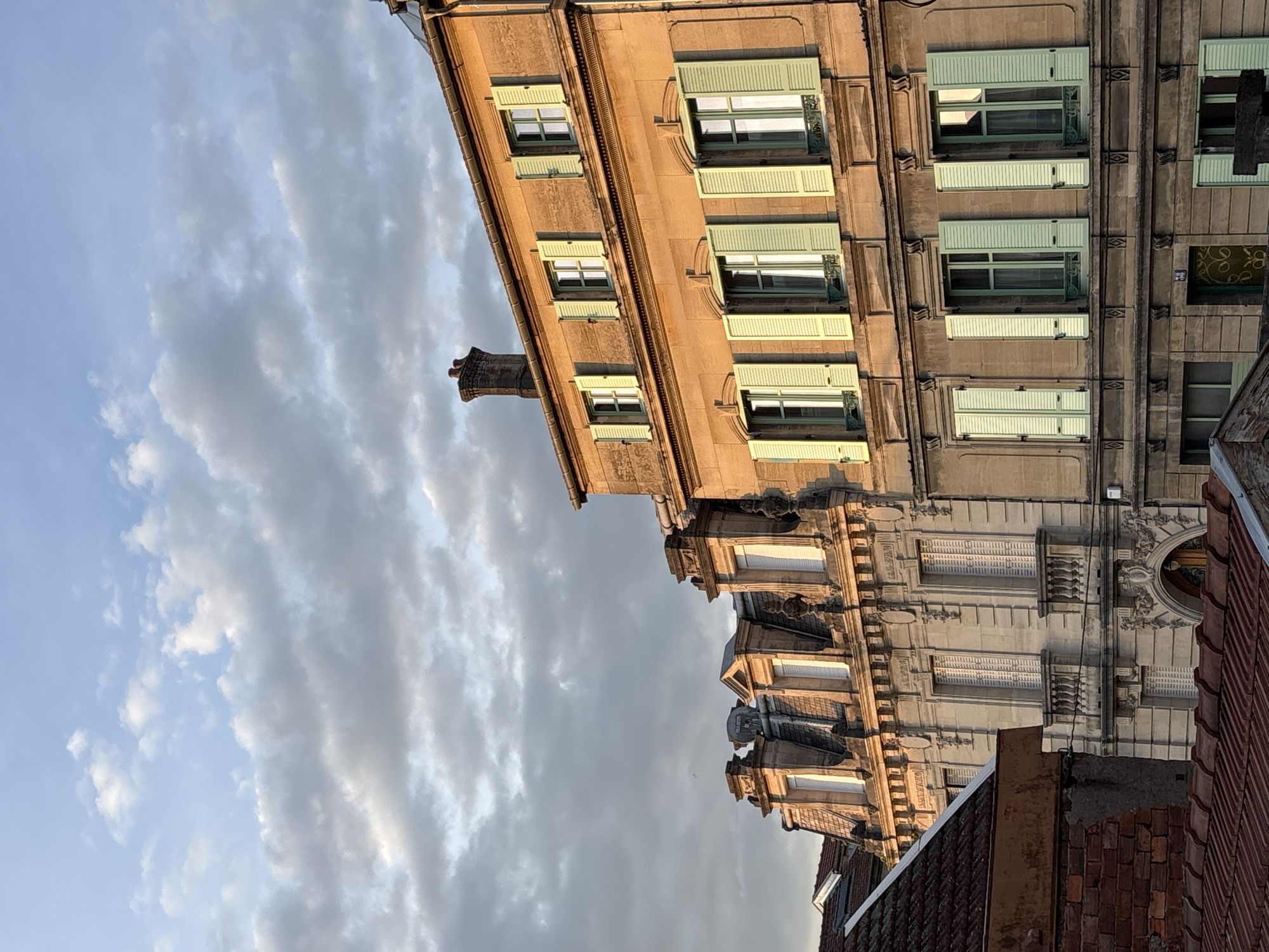 The image shows a building in Bar-le-Duc, France, viewed from below at an angle. The sky is cloudy. The building's facade features rows of windows with light green shutters. The building's architecture shows intricate details and molding. Part of a red-tiled roof is visible in the foreground.