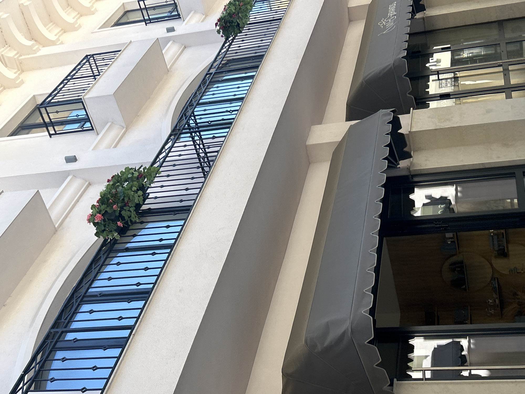 The image shows an upward angle view of a building facade. The building is light-colored, possibly white or cream, and features multiple windows. Some windows are enclosed with black iron railing and adorned with potted plants. Awnings, also dark-colored, possibly grey or black, are visible over other windows. The architectural style appears modern with classical influences, incorporating elements like rounded arches. The sky is visible through the windows. The scene appears to be an urban environment, but the specific location is unclear.
