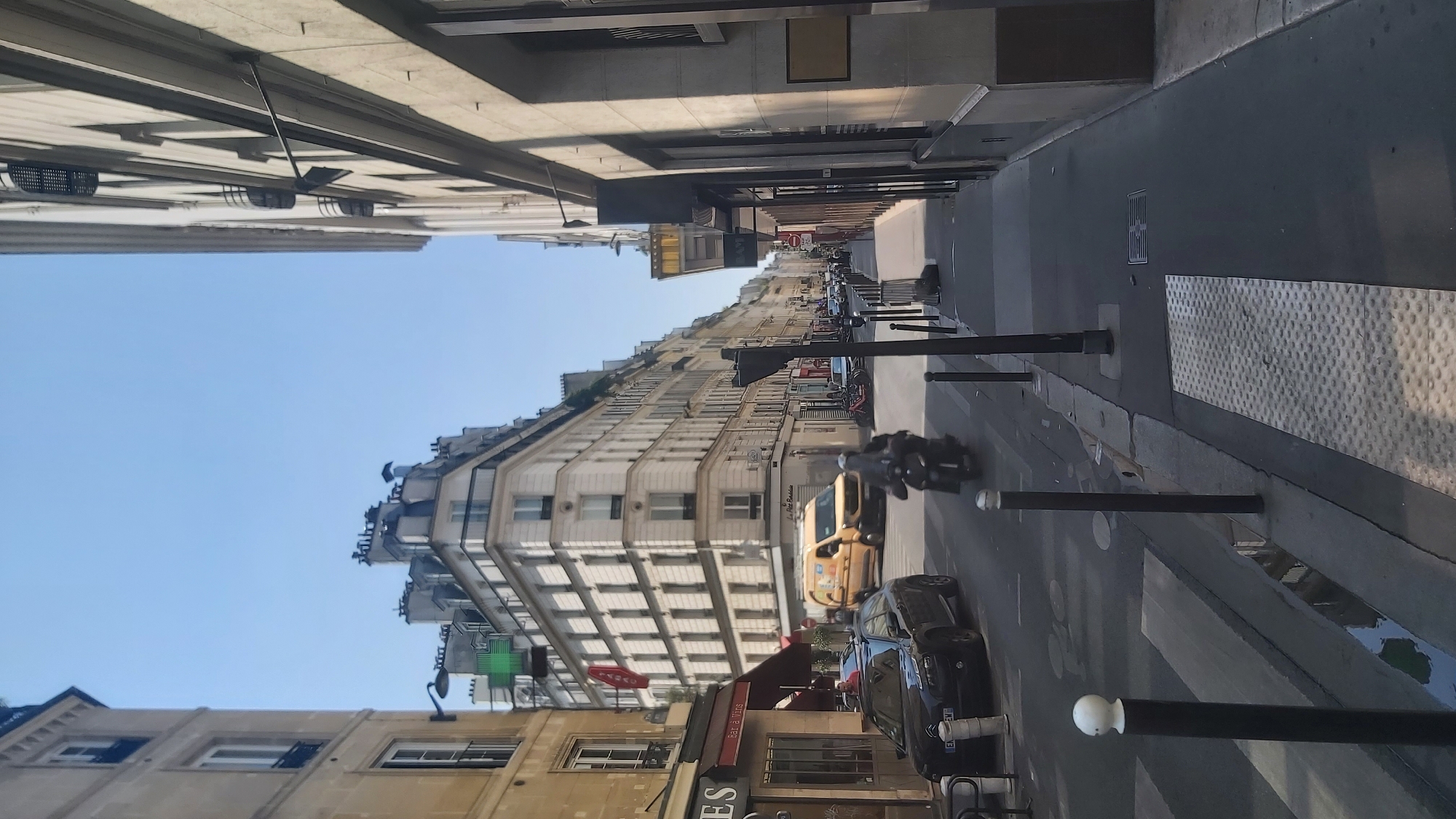 The image depicts a street scene in Paris, France, on a clear, sunny day. The perspective is from a pedestrian's eye level, looking down a street lined with tall, multi-story buildings. The architecture is characteristic of Parisian buildings, with stone facades, numerous windows, and decorative elements.
On the left side of the street, there are shops at ground level, one with an awning and signage that includes "ES" and "bar à Vins". A black car is parked partially on the street and sidewalk. Further down the street on the left, a yellow van is visible.
The right side of the street features a sidewalk with more shops or businesses at ground level. The buildings on the right appear to be slightly more modern in design, with some darker glass facades.
In the middle of the street, a person is riding a scooter, with their body blurred due to motion. Another car is visible further down the street, along with several parked bicycles. The street is paved with asphalt and cobblestones, and there is a pedestrian crossing marked on the road.
Streetlights and bollards are present along the sidewalk and street. The sky is a clear blue, indicating fair weather. The overall impression is of a typical urban street in Paris during daylight hours.