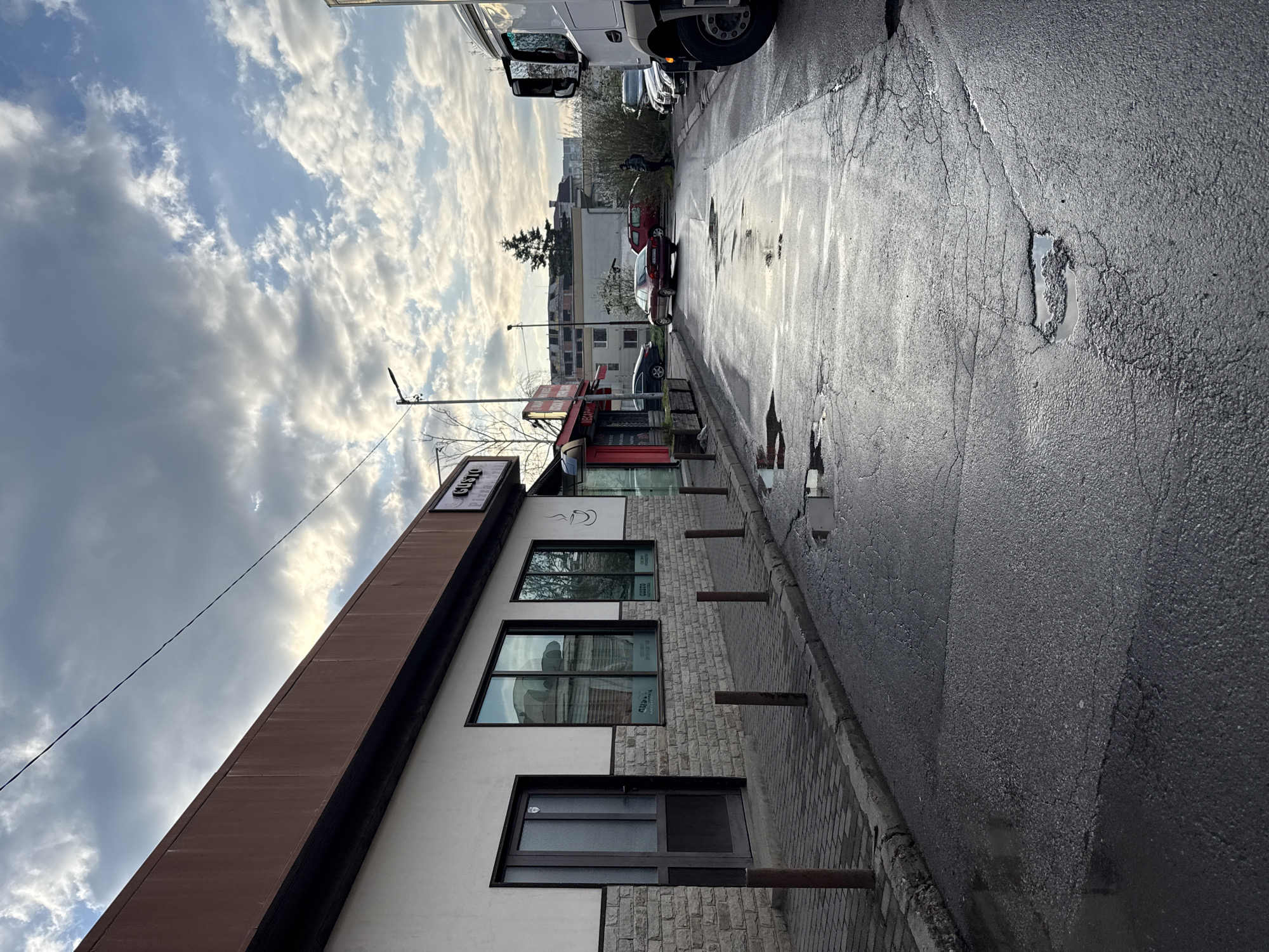 The image depicts a street scene in Sofia, Bulgaria. The street is wet, suggesting recent rainfall, and shows signs of wear with cracks and potholes. A building with a sign that appears to read "GUSTO" is visible on the left. The building has a white facade and three windows. A row of metal posts lines the sidewalk in front of the building. Cars are parked along the right side of the street. The sky is cloudy with patches of blue. The camera is angled slightly upwards.