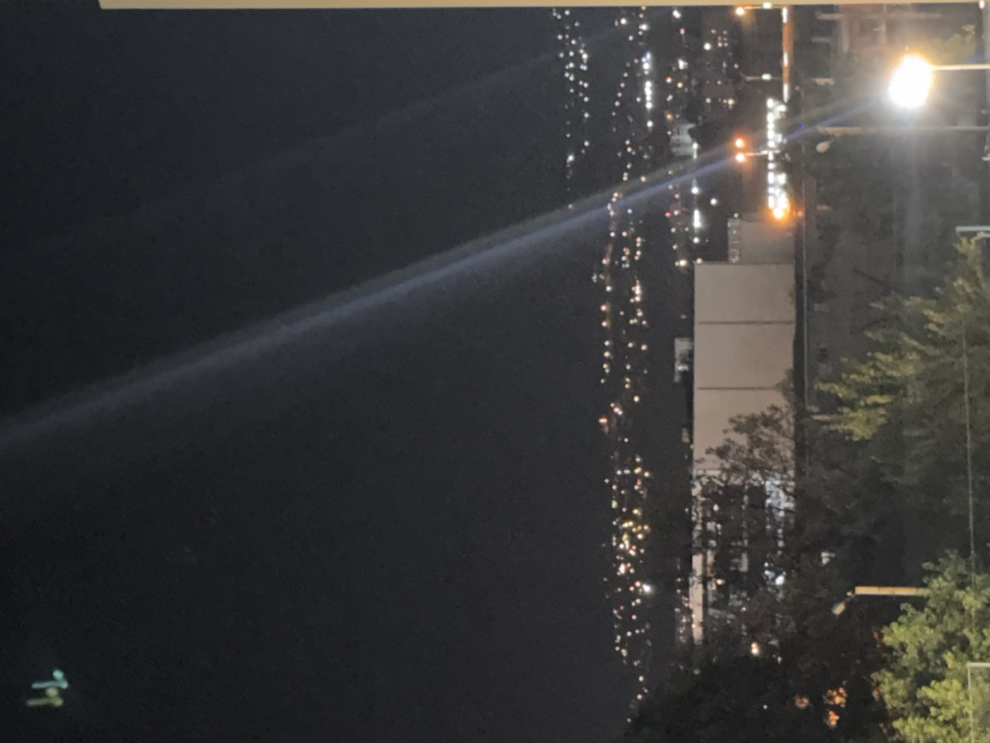 The image shows a night view of the city of Tbilisi, Georgia. In the upper left corner, there is a distant, small, bright object in the sky, possibly an aircraft or drone. The background consists of a city skyline with numerous lights, indicating an urban area at night, nestled amongst hills. A bright beam of light, possibly from a searchlight or stage lighting, cuts diagonally across the image from the top left to the bottom right. In the foreground and middle ground, there are trees and some buildings. One prominent building is large, rectangular, and white. There are also streetlights visible, one of which is bright and illuminated in the bottom right of the frame, and another further back with an orange hue. The overall scene depicts a nighttime urban landscape.