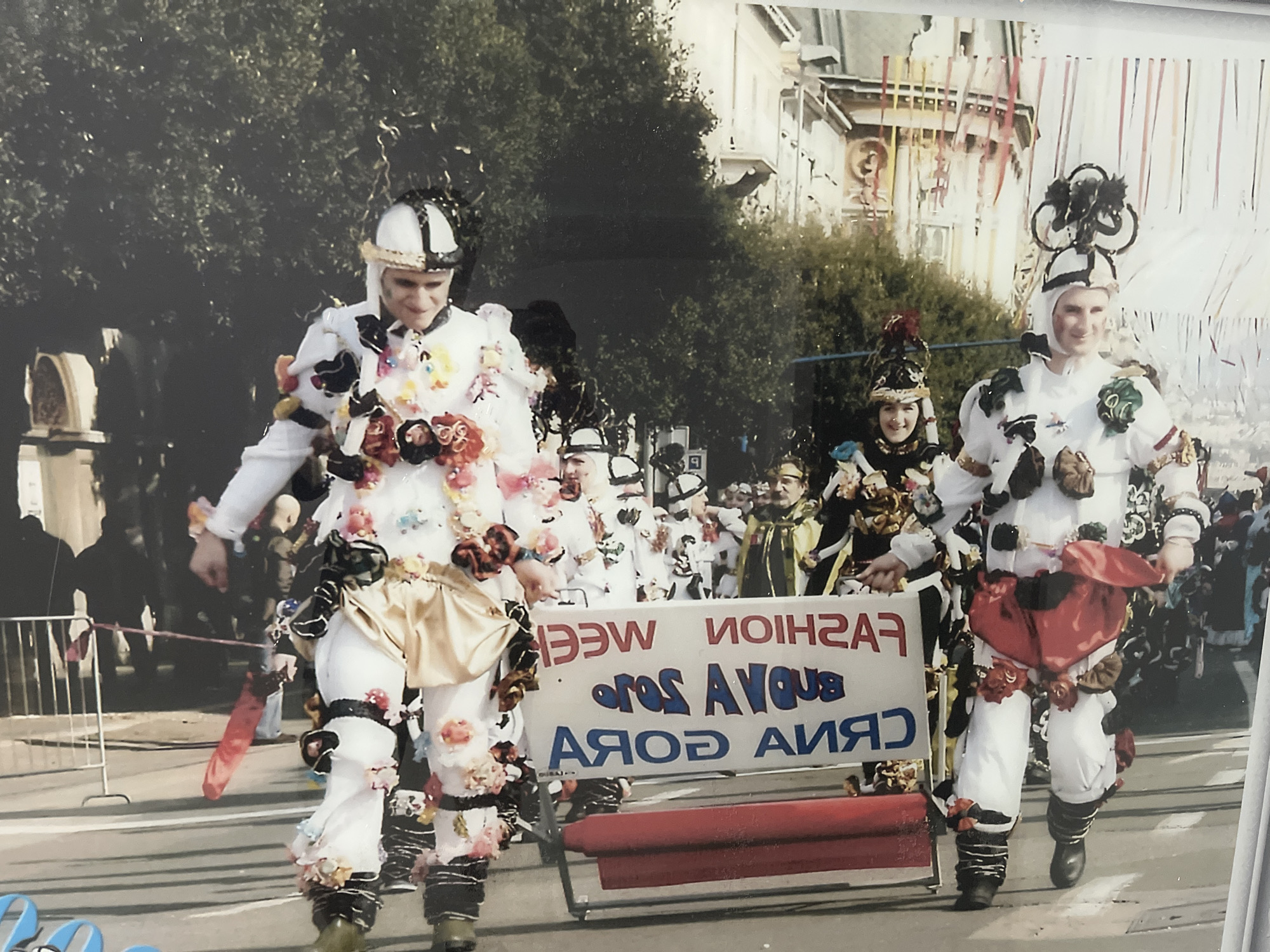 This image depicts a street parade in Budva, Montenegro. The parade features individuals in elaborate costumes, likely participating in a festival or carnival. The central figures are dressed in white, full-body costumes adorned with colorful floral embellishments and headwear. The costume on the right includes a large red heart-shaped accessory worn as a cape. Behind the costumed participants, a banner is visible with text that appears to be reversed and possibly in Cyrillic or a similar alphabet. The text reads "FASHION WEEK BUENA GORA". In the background, buildings with decorated facades and trees suggest an urban setting. People are lined up along the street, watching the parade, and some are holding what appear to be cameras. A metal barrier is present, separating the parade route from the spectators. The overall atmosphere suggests a celebratory event taking place outdoors during daylight.