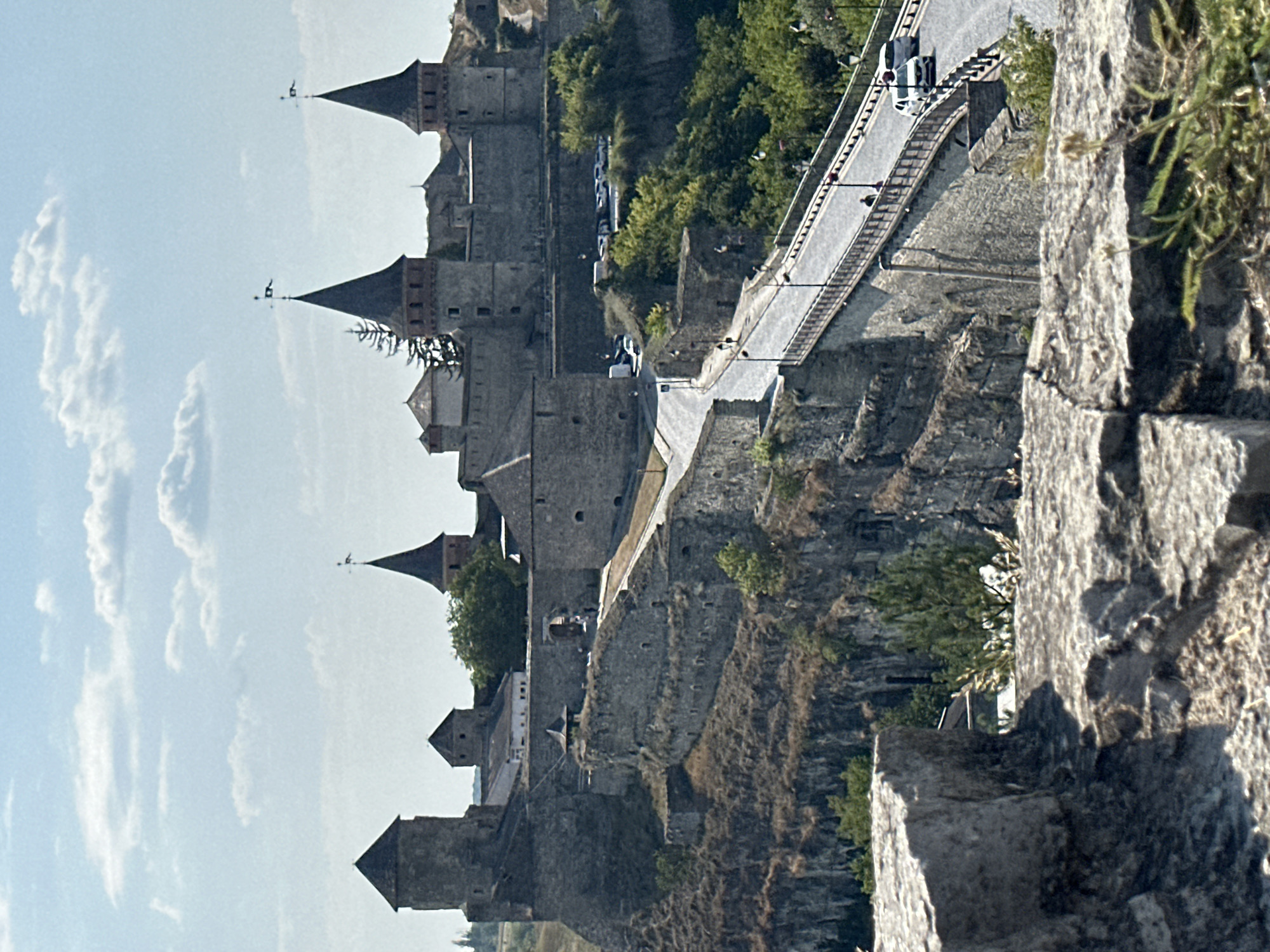 The image shows the Kamianets-Podilskyi Castle in Ukraine. The castle is situated on a rocky promontory, with a deep canyon visible in the foreground. A bridge spans the canyon, leading to the castle entrance. Cars and pedestrians are present on the bridge and the road approaching the castle. The sky is blue with scattered clouds. The castle itself is a large stone structure with several towers and fortifications. The surrounding landscape is characterized by steep cliffs and lush greenery.