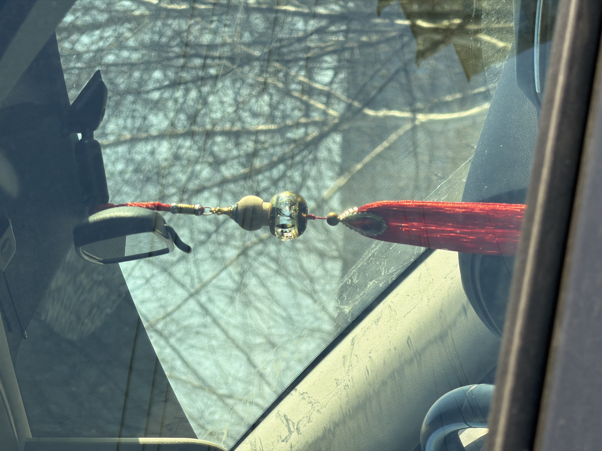 The image depicts an object hanging from a car's rearview mirror. The object appears to be a decorative tassel with wooden and glass beads. The background shows a view through the car's windshield, possibly depicting trees. The interior of the car is visible, including the dashboard and parts of the steering wheel. There is no information present indicating that this image was taken in Izhevsk, Russia.