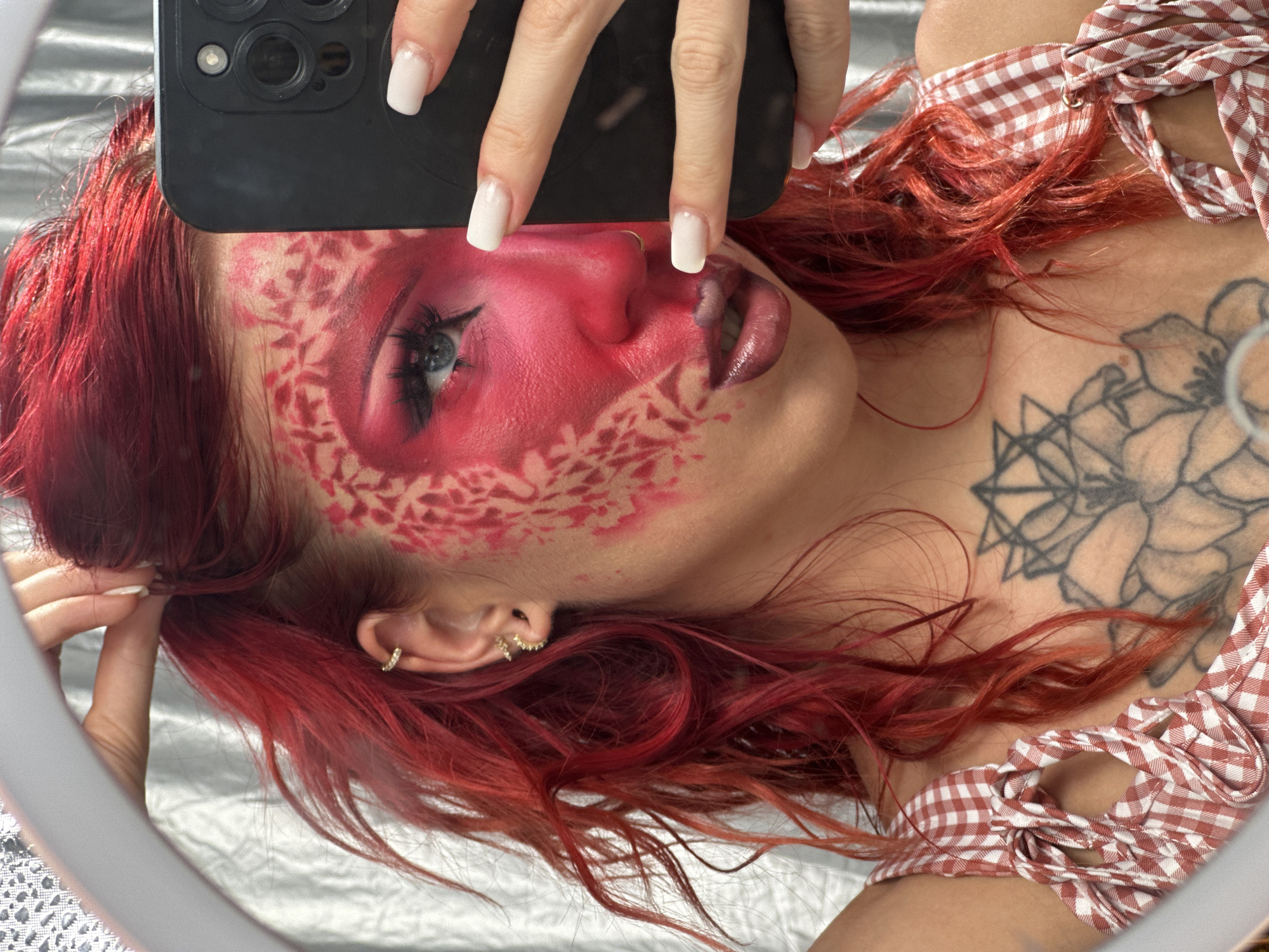 A close-up, high-angle shot captures a woman with bright red hair, styled loosely around her face. She is taking a selfie in a ring light with a black smartphone. Her face features dramatic red face paint, with a patterned design in a lighter red or pink around her left eye and cheek. Her makeup includes bold eyeliner, lashes, and dark lipstick. She is wearing a red and white gingham top with tie straps. A large, intricate black tattoo is visible on her upper chest. The background appears to be a textured silver curtain. The scene does not contain any identifiable elements indicating a location in North Kuta, Indonesia.