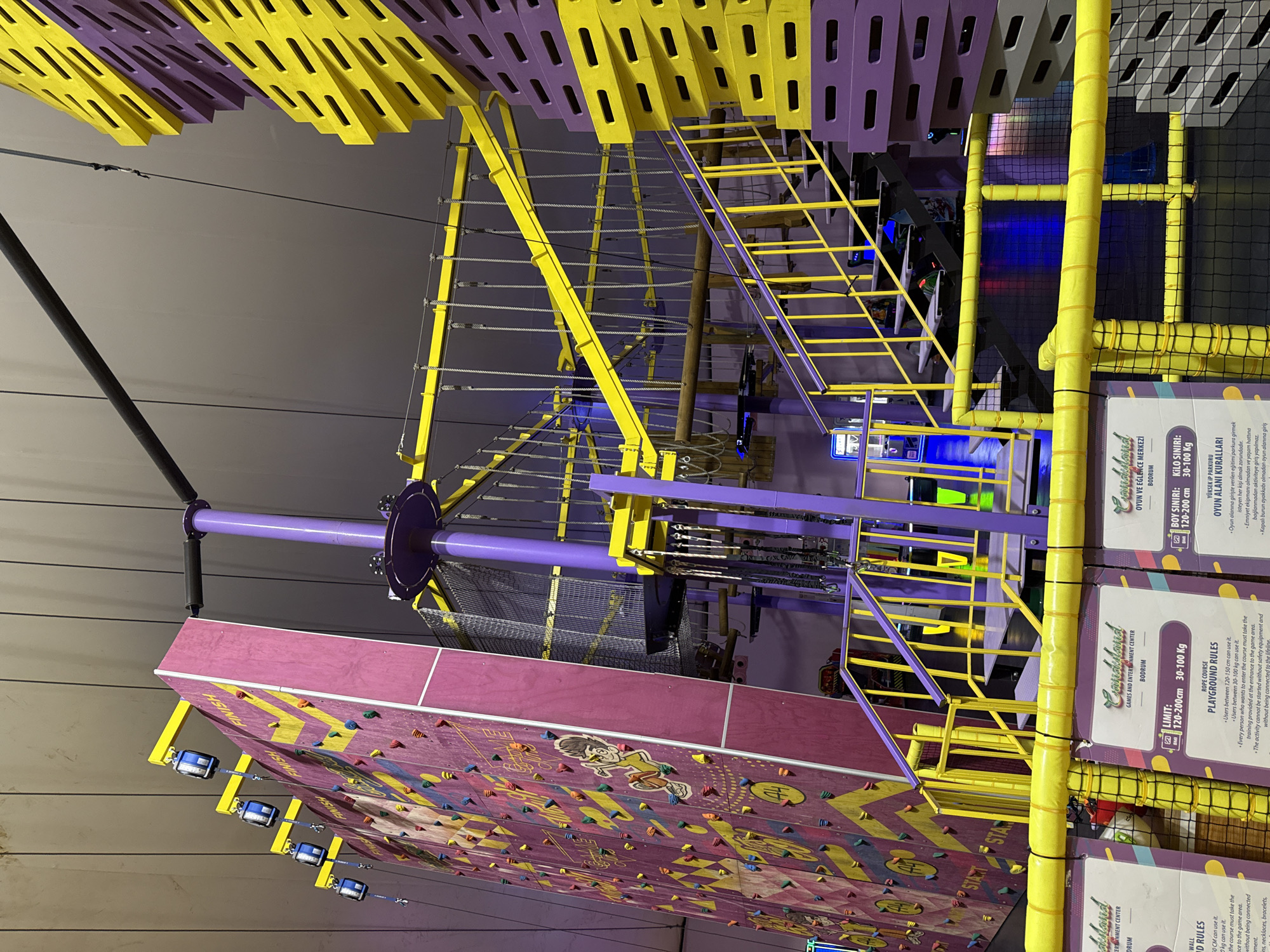 The image depicts an indoor adventure park in Bodrum, Turkey, likely "Candyland Games and Entertainment Center." The foreground features yellow and purple informational signs with text in Turkish, detailing playground rules and weight/height limits for activities such as a rope course. A climbing wall, predominantly pink with yellow diagonal stripes and colorful holds, dominates the left side of the frame. Suspended from the ceiling are various elements of a rope course, including cables, netting, and wooden platforms, all supported by purple poles and yellow metal beams. To the right, a tall, stacked structure made of interlocking yellow and purple rectangular blocks rises upwards, suggesting a climbing or obstacle feature. A yellow staircase with purple railings leads up towards the rope course. The environment appears to be a brightly lit indoor space with a gray ceiling. There are no people visible in the frame.