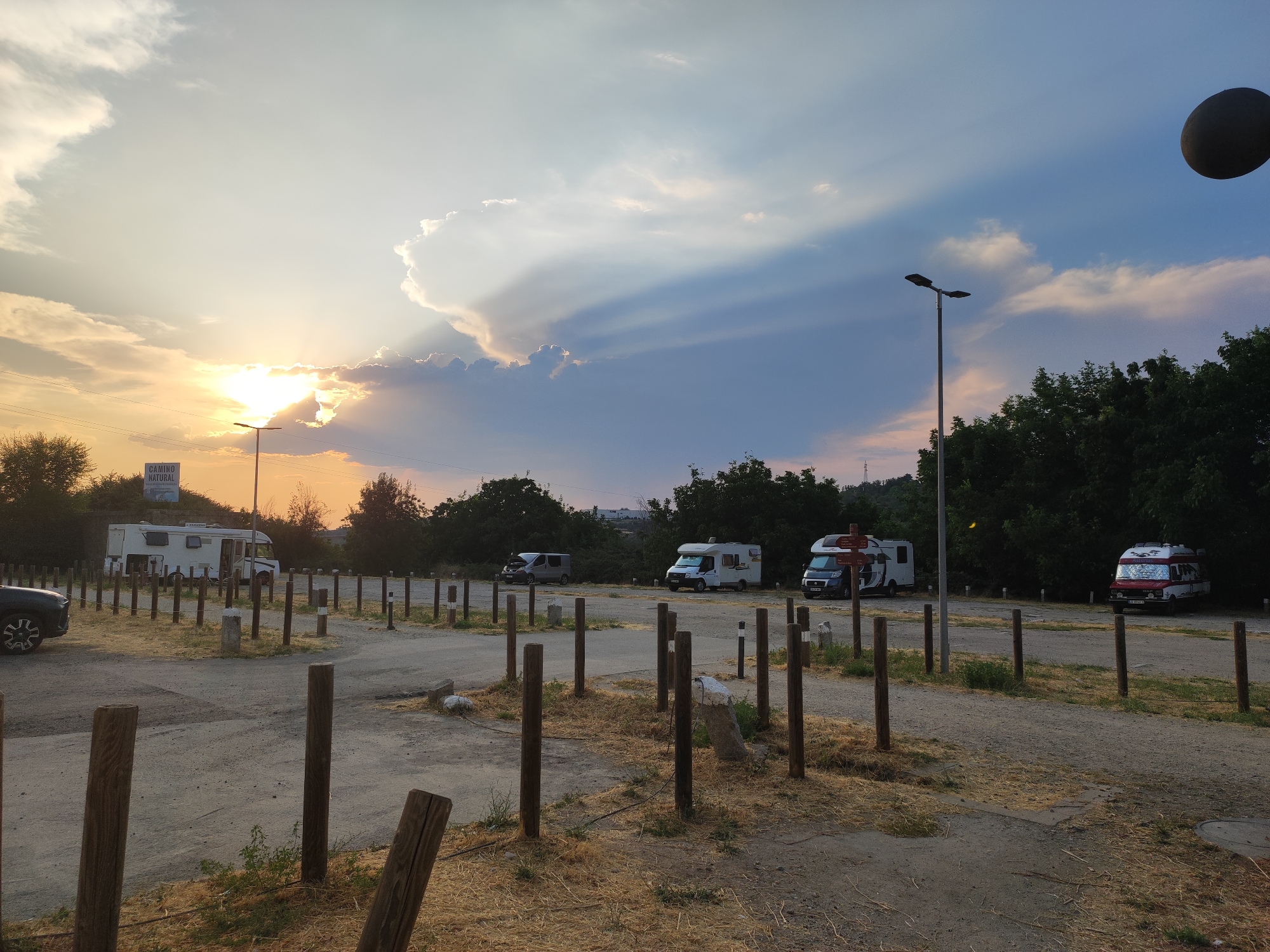 The image depicts a campsite at dusk, likely located in Béjar, Spain. The sun is setting, casting a warm glow and dramatic light rays through the clouds. Several recreational vehicles (RVs) are parked in an open, dusty area marked by wooden posts. In the background, a line of trees forms a dense border. A sign that reads "CAMINO NATURAL" is visible on a pole, indicating a natural path or trail, which is characteristic of rural or semi-rural Spanish landscapes. The overall atmosphere is serene, with the fading sunlight creating a peaceful and tranquil scene. There are no people or active interactions visible. The ground is covered in gravel and dry grass, typical of a campsite setting. The sky is a mix of blues, grays, and oranges due to the sunset.