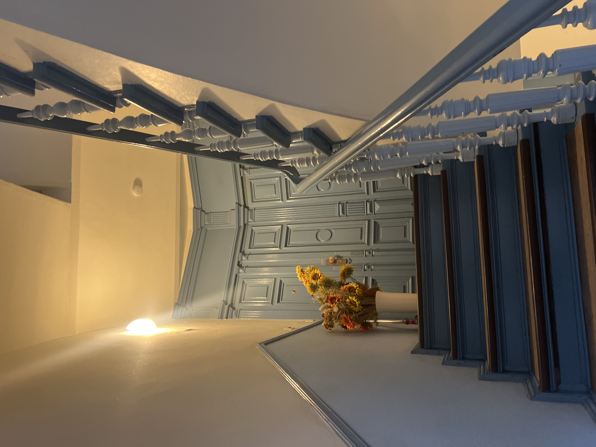 The image shows an indoor staircase with blue painted steps and wooden treads. The banister is also painted blue and has decorative spindles. To the left of the staircase, there is a wall with a light fixture. A white vase with a bouquet of sunflowers and other flowers is placed on the top step. In the background, there is a large, light blue double door with decorative paneling. The scene appears to be in an apartment building in Berlin, Germany, likely in a residential area. The lighting suggests it is either evening or the interior lighting is on. There are no people or animals visible in the image.