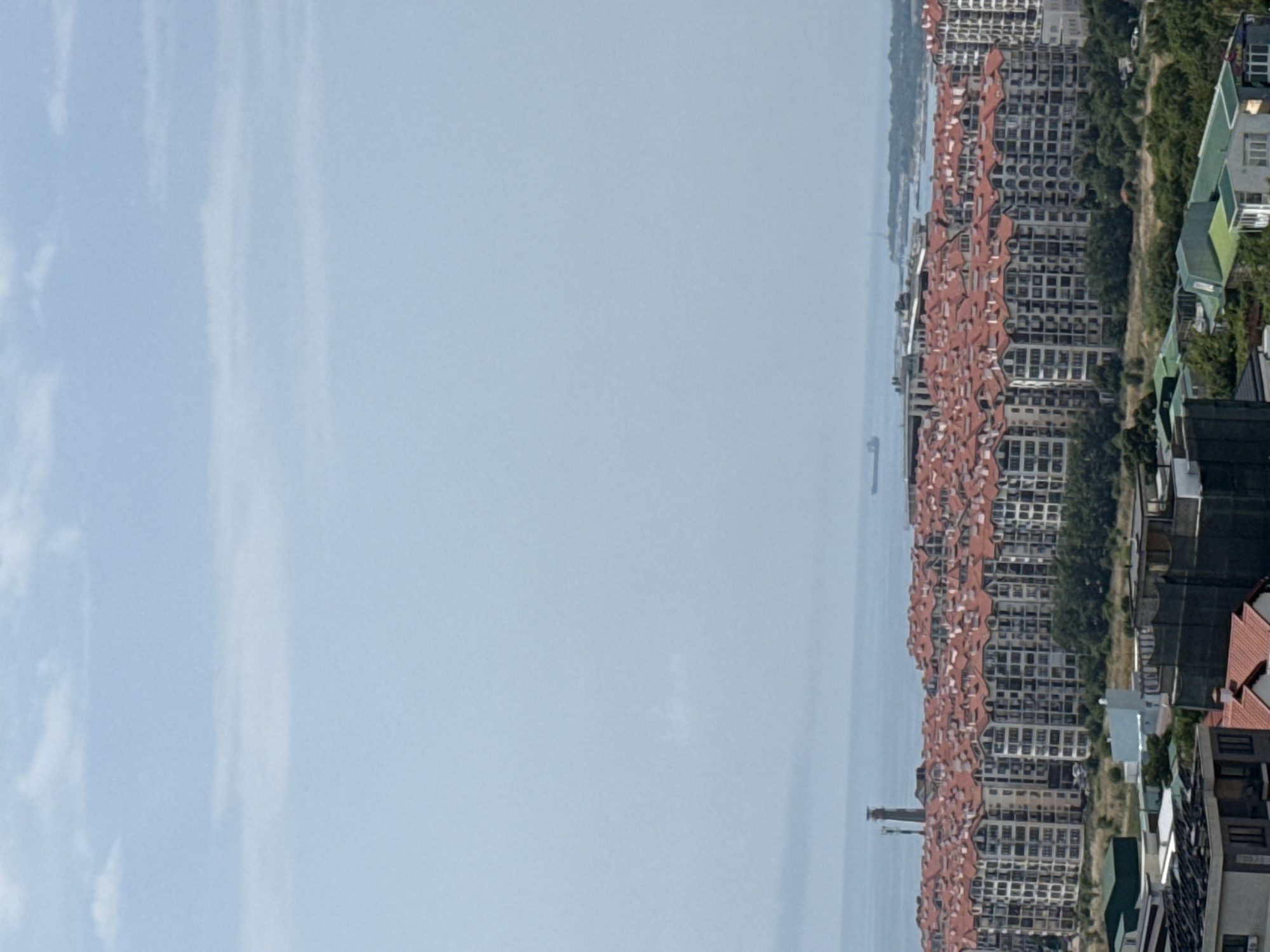 The image is an aerial view of a residential area in Gelendzhik, Russia. In the foreground, there are several buildings with pitched, red-tiled roofs, some with visible construction or renovation. Behind these, a large complex of apartment buildings with identical red roofs dominates the mid-ground. These buildings have a consistent architectural style, featuring multiple balconies and white or light-colored facades.
Beyond the apartment complex, a body of water, likely the Black Sea, stretches to the horizon. A ship is visible on the water in the distance, and what appears to be a lighthouse or tower is situated on the coastline. The sky is overcast with a hazy, pale blue hue. The overall impression is of a densely populated coastal urban or suburban environment.