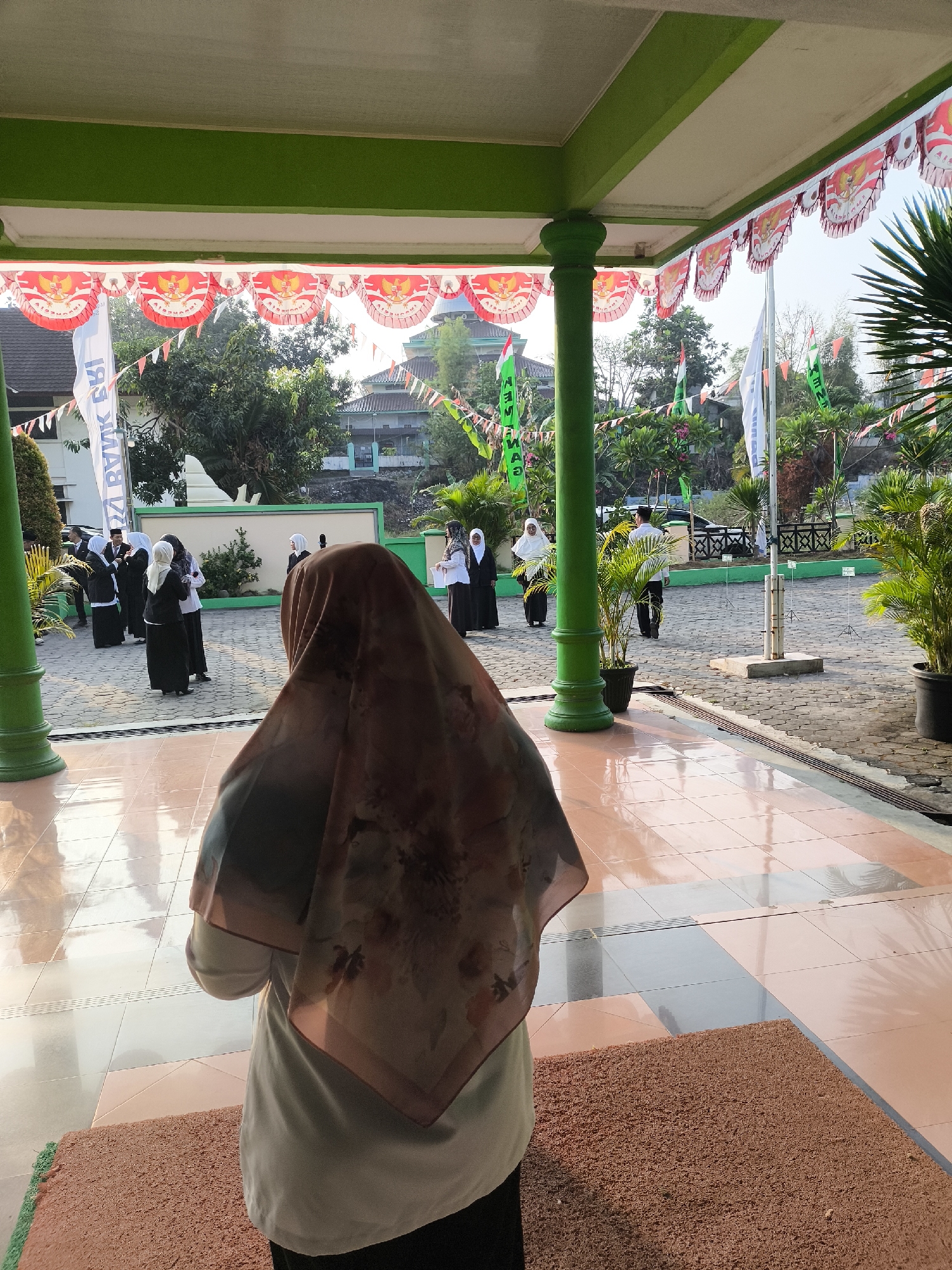 In the city of Cilegon, Indonesia, a group of people, mostly women wearing hijabs and dark uniforms, are gathered in an outdoor courtyard. The courtyard has a tiled floor and is decorated with red and white pennant banners, some featuring the Garuda Pancasila emblem. Several flags, including a white one and some green ones with text, are visible. In the foreground, a woman wearing a patterned hijab and a light-colored top stands facing away from the camera, looking towards the gathering. The scene appears to be taking place on a sunny day, with shadows cast on the ground. The architectural elements suggest a public building or institution, possibly a school or government facility, with green pillars supporting an overhang. Lush green plants in pots are also present around the perimeter.