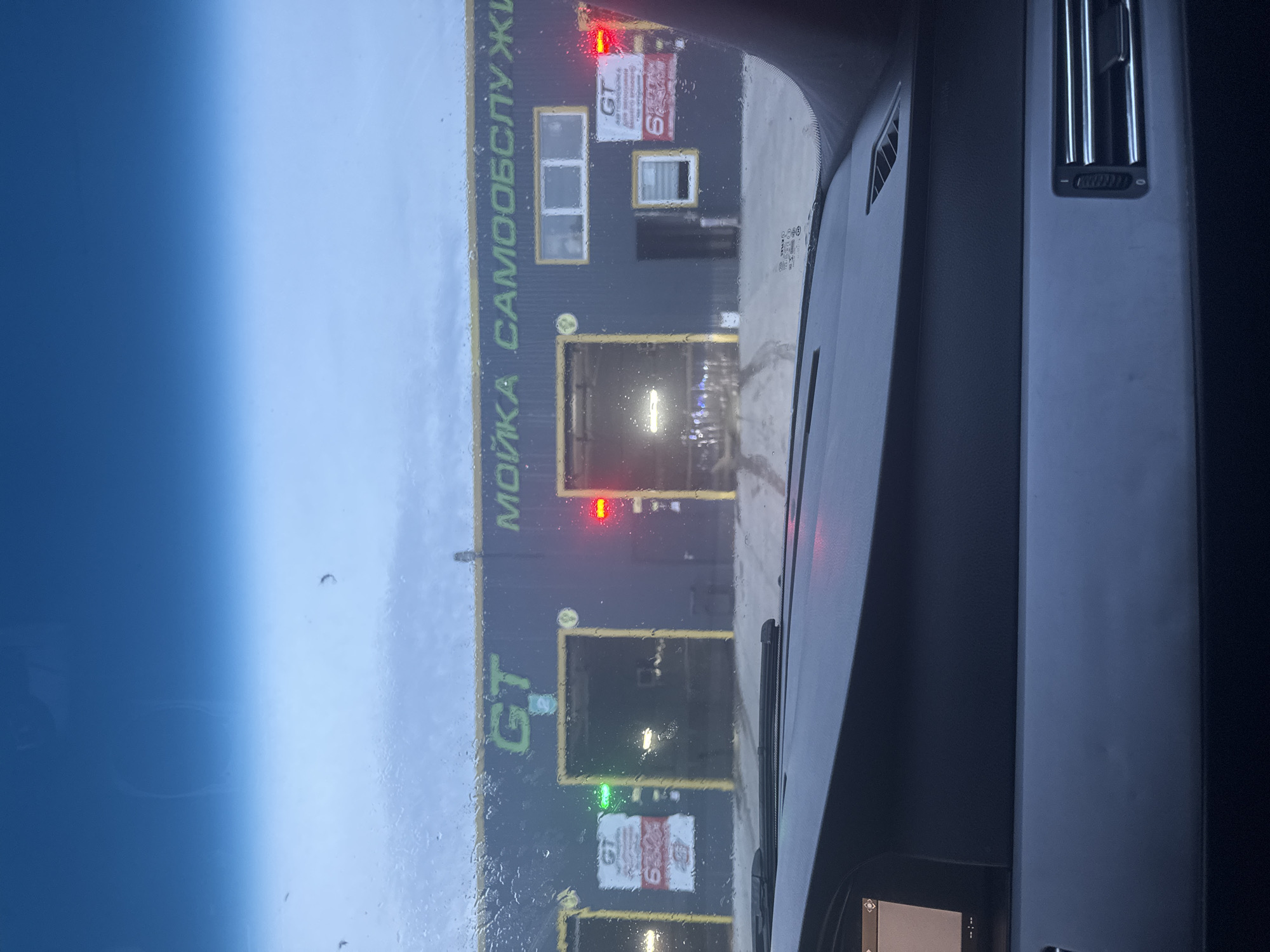 The image, taken from inside a car, shows the "GT" car wash facility in the city of Chaikovsky, Russia. The building is dark grey with green lettering in Russian. Multiple garage doors are visible, with some showing red and green lights indicating the status of each bay. There are raindrops on the car's windshield. The car's dashboard and air vents are visible in the interior.