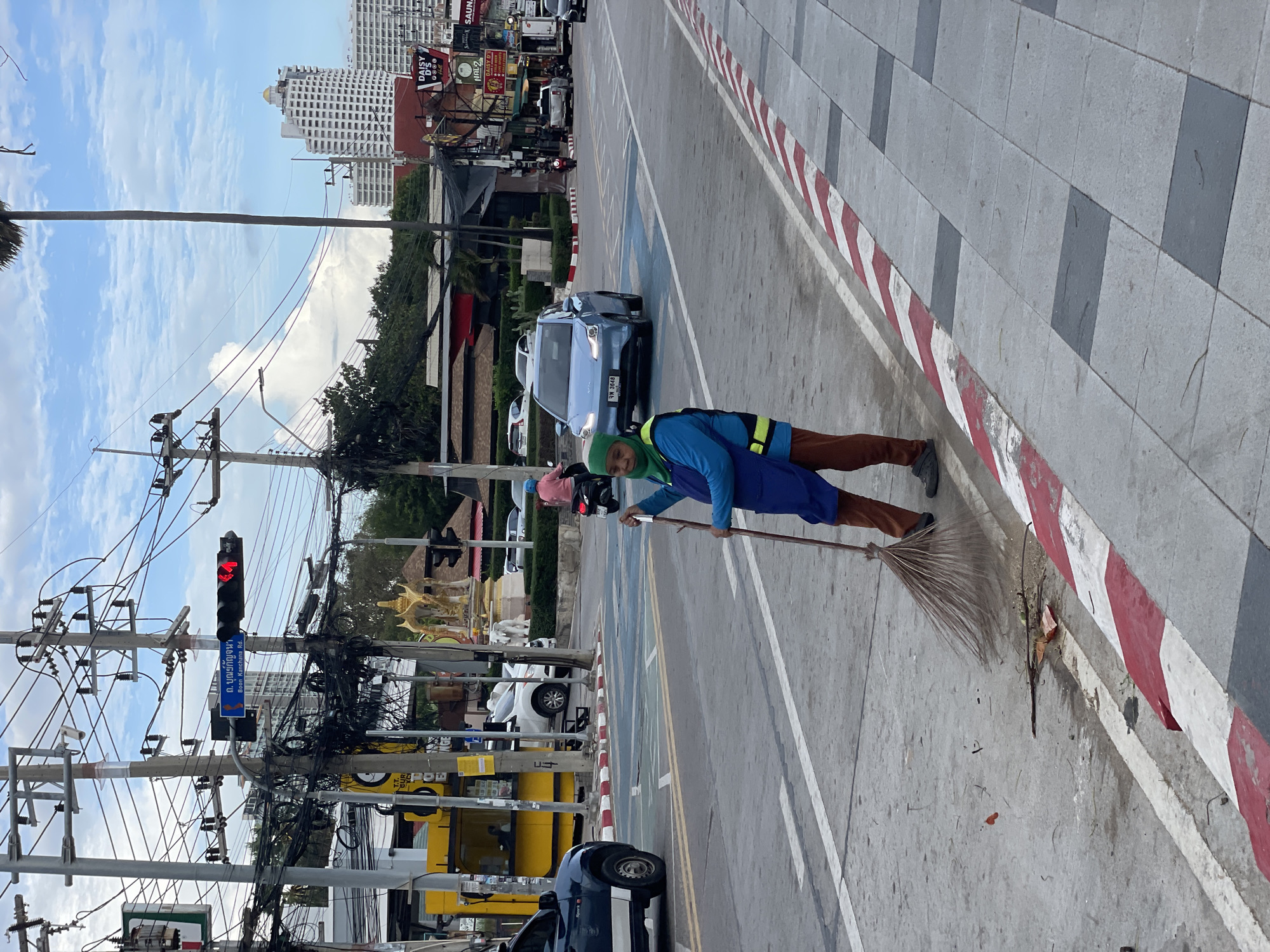 A woman with a green headscarf and blue apron sweeps the street in Pattaya City, Thailand. She is standing on the side of a busy road, with cars and motorcycles passing by. The woman is holding a broom and appears to be cleaning up debris from the sidewalk. The road is lined with shops and buildings, and there are traffic lights and power lines overhead. The sky is cloudy, and the weather appears to be overcast.