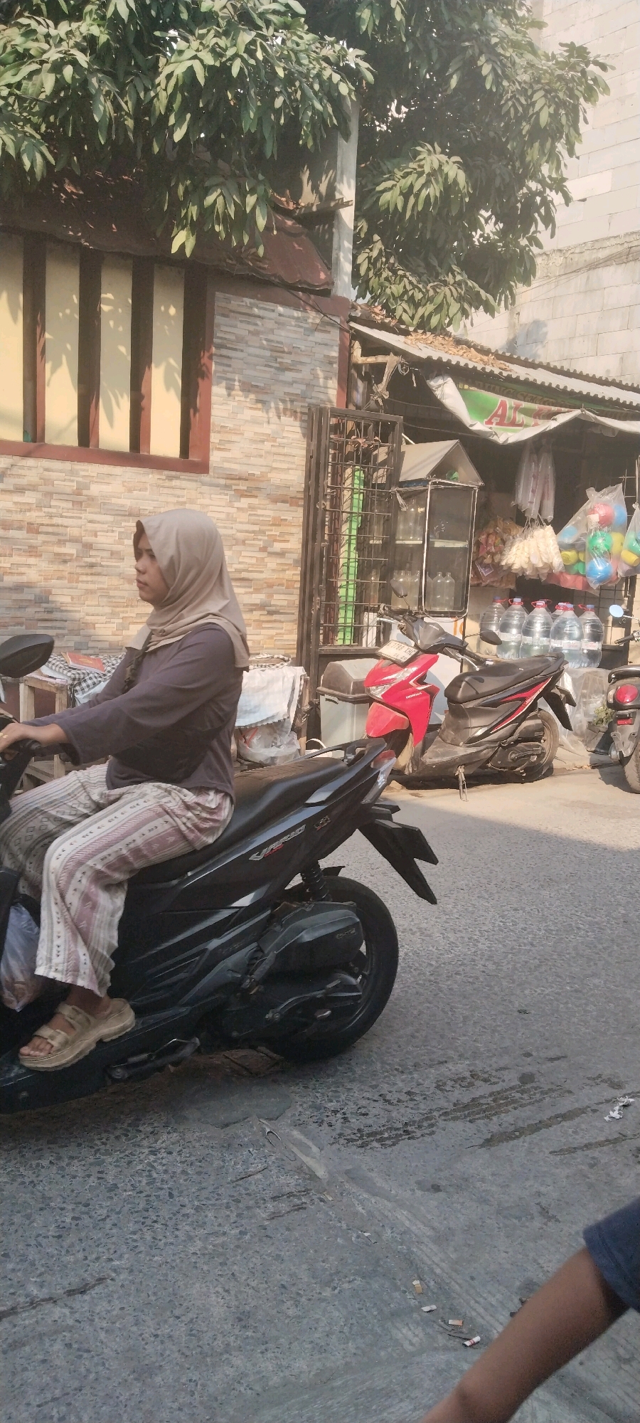 A woman wearing a light brown hijab and striped pants is riding a black motorcycle on a paved street in front of a building with a textured wall. She is looking to her left and has her right hand on the handlebars. A red motorcycle is parked behind her. To the right of the motorcycles, there is a small stall with various items displayed, including bottled water, bags of snacks, and colorful balls. Overhead, a large tree with green leaves casts shadows on the scene. The environment appears to be a street in a city, likely in West Jakarta, Indonesia, given the context.